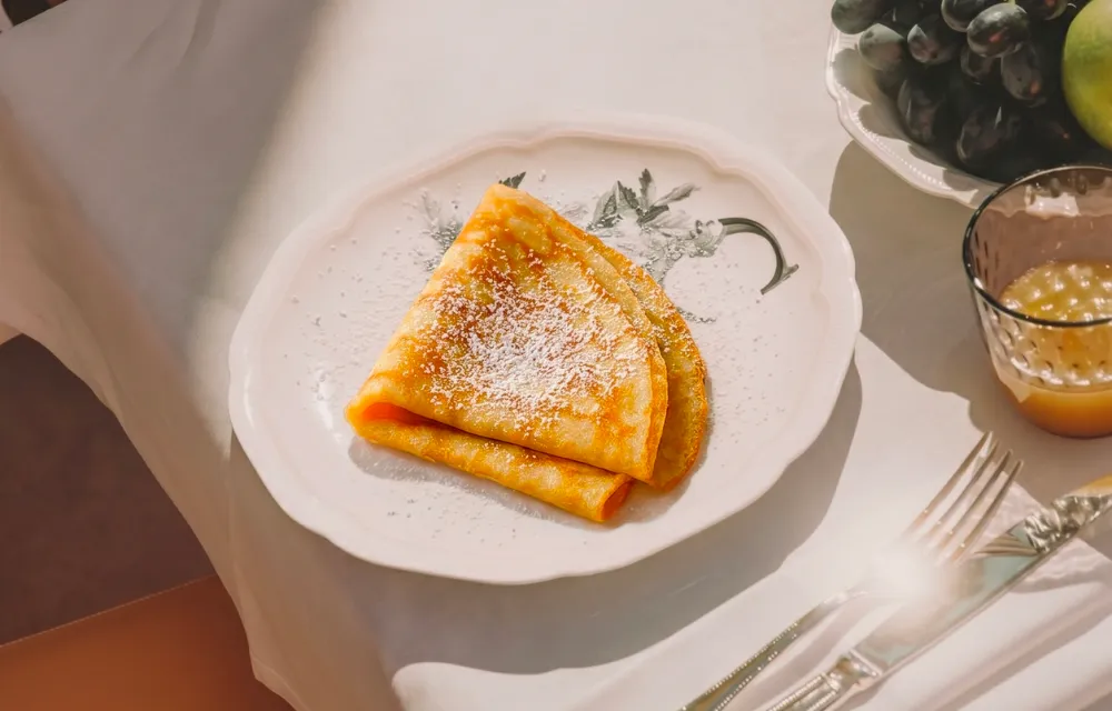 Crêpes dorées saupoudrées de sucre glace sur une assiette élégante à la Villa Colette, servies pour le petit-déjeuner avec des fruits frais.