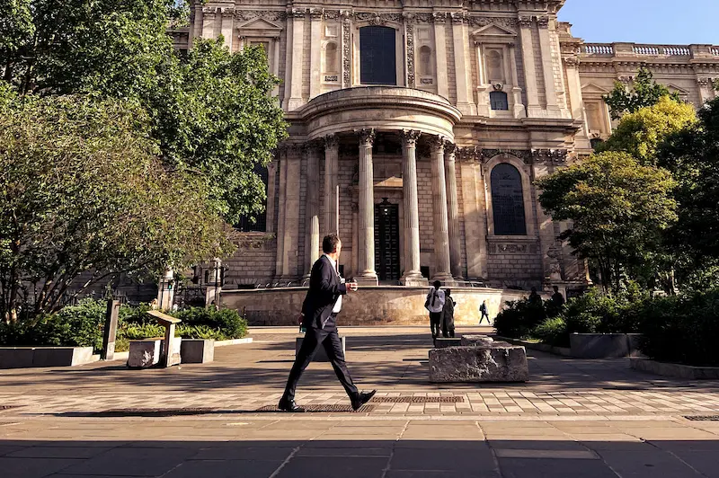 Een man in een donker pak loopt over een plein met bomen voor een groot klassiek gebouw met zuilen.