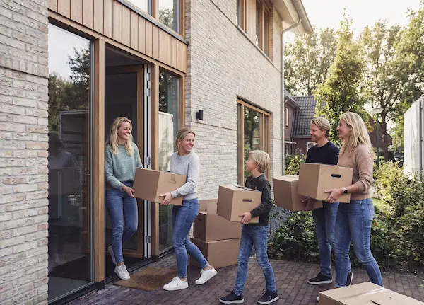 Vier personen dragen dozen terwijl ze een huis binnenlopen in een zonnige woonwijk.