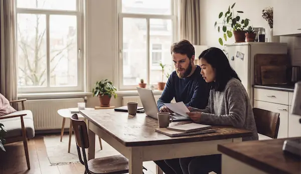 Twee personen zitten aan een houten tafel in een lichte kamer, ze bekijken samen documenten terwijl ze een laptop gebruiken.
