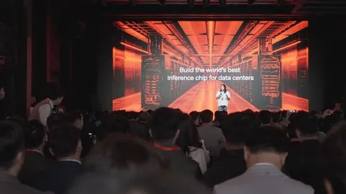 Speaker presenting on stage in front of a large screen showing data center equipment and text about inference chips.