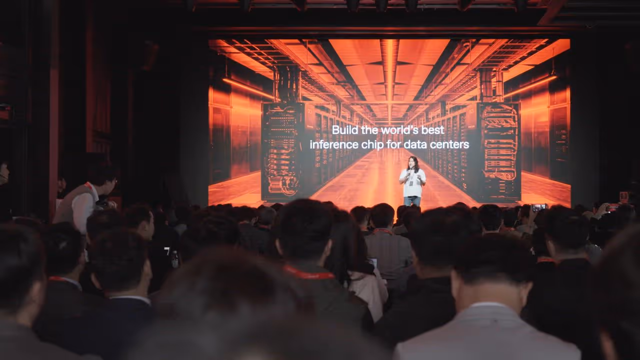 Speaker presenting on stage in front of a large screen showing data center equipment and text about inference chips.
