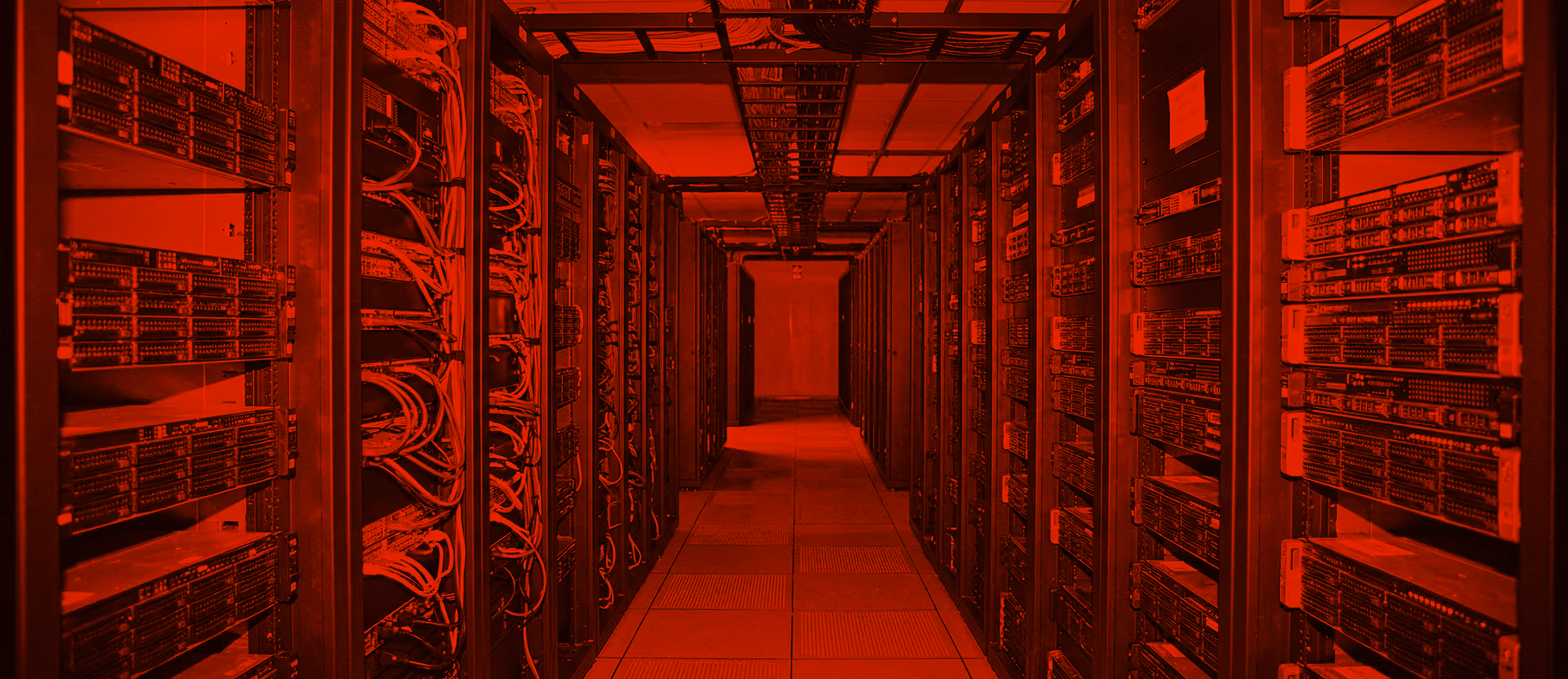 A narrow aisle in a data center with tall racks of servers and network cables on both sides, bathed in red lighting.