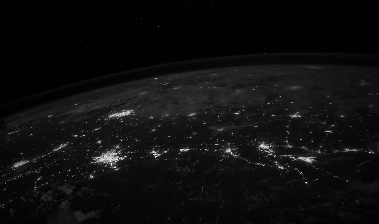 View of Earth's night side from space showing illuminated city clusters and roads across a large land area under a starry sky.