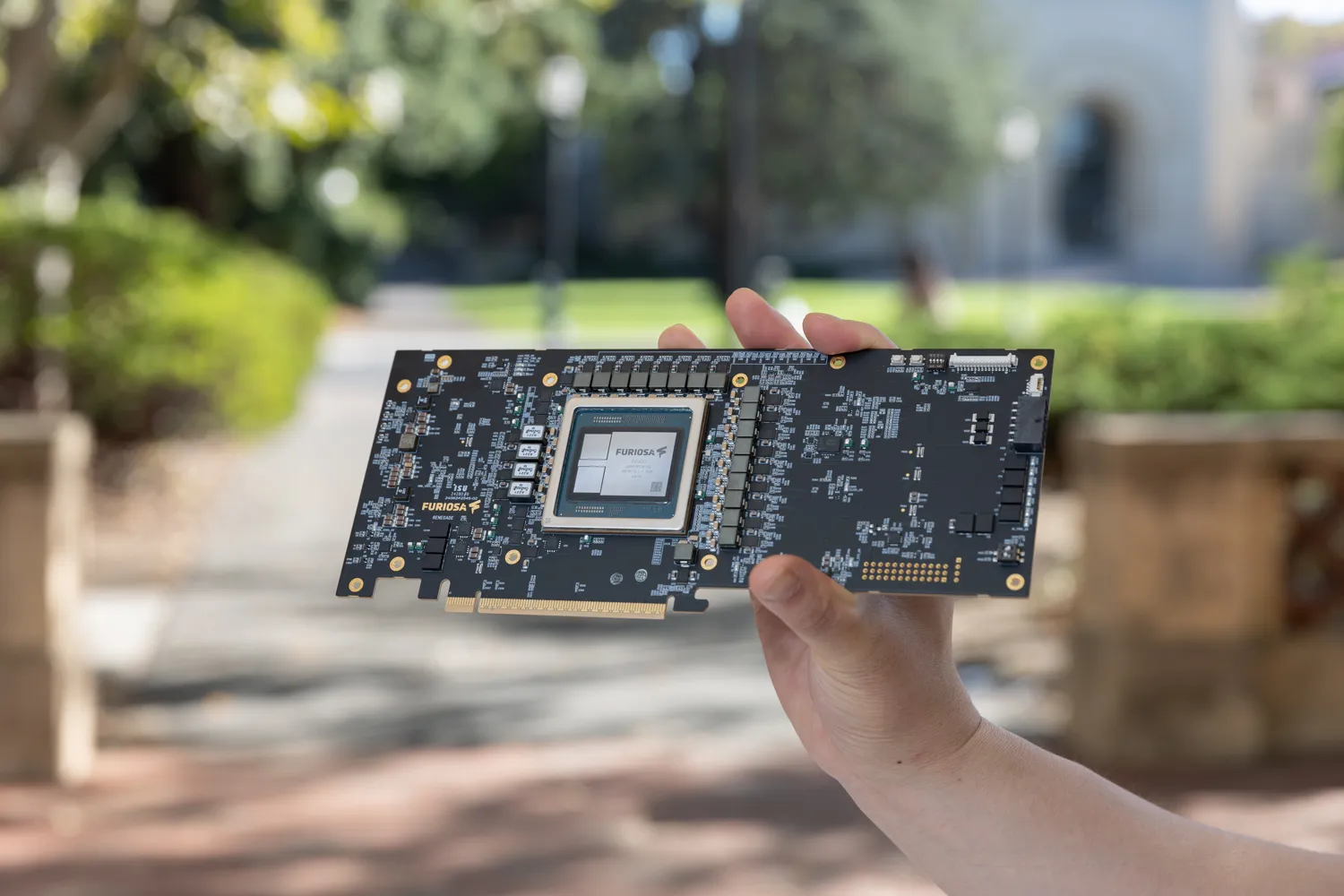 A hand holding a FuriosaAI accelerator card outdoors, showing the exposed circuit board and central Furiosa-branded chip, with electronic components and connectors visible against a blurred garden and walkway background.