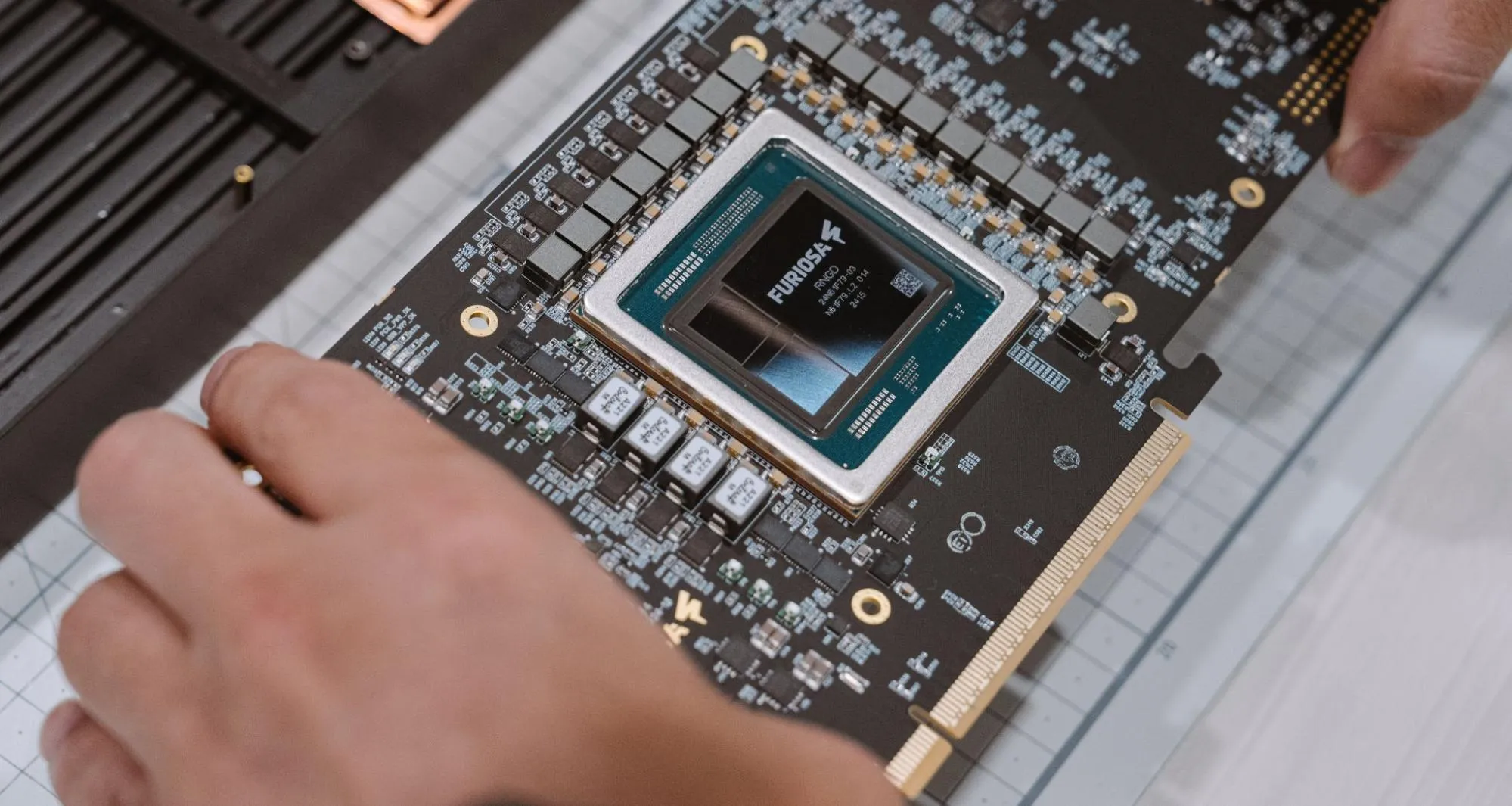 Close-up of a FuriosaAI accelerator card on a work surface, showing the central Furiosa-branded chip surrounded by dense circuitry and power components, with two hands carefully positioning or inspecting the board during hardware assembly or testing.