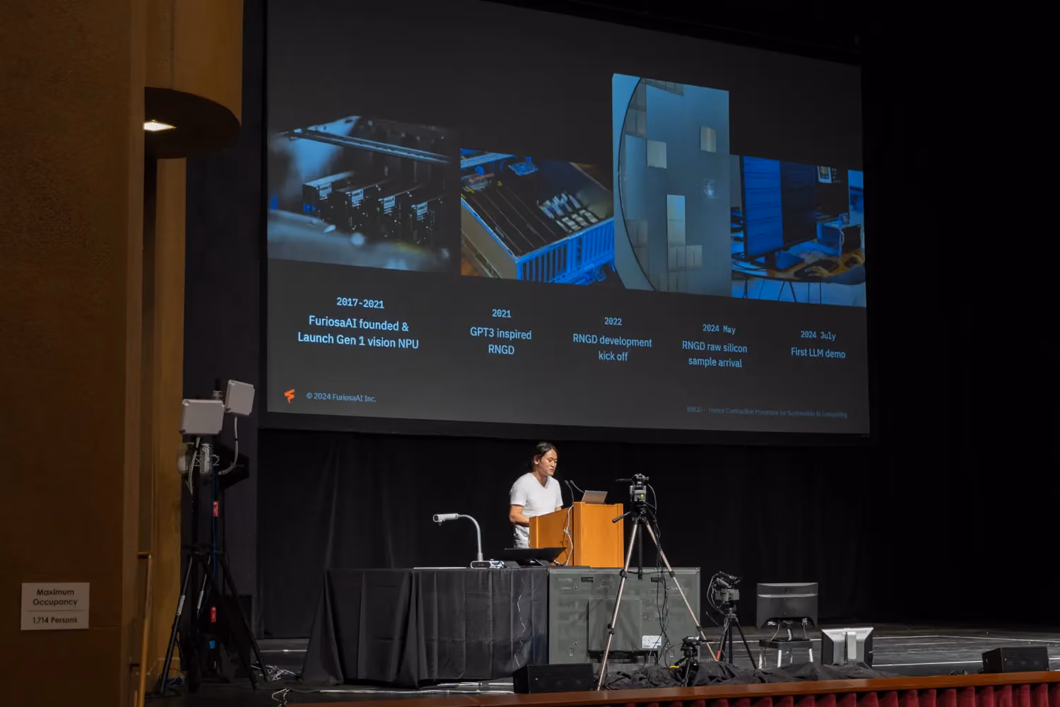 Wide-angle view of a speaker presenting on stage at a large auditorium during a tech conference, with a projected slide behind them showing FuriosaAI’s product timeline, milestones, and hardware images related to AI accelerator development.