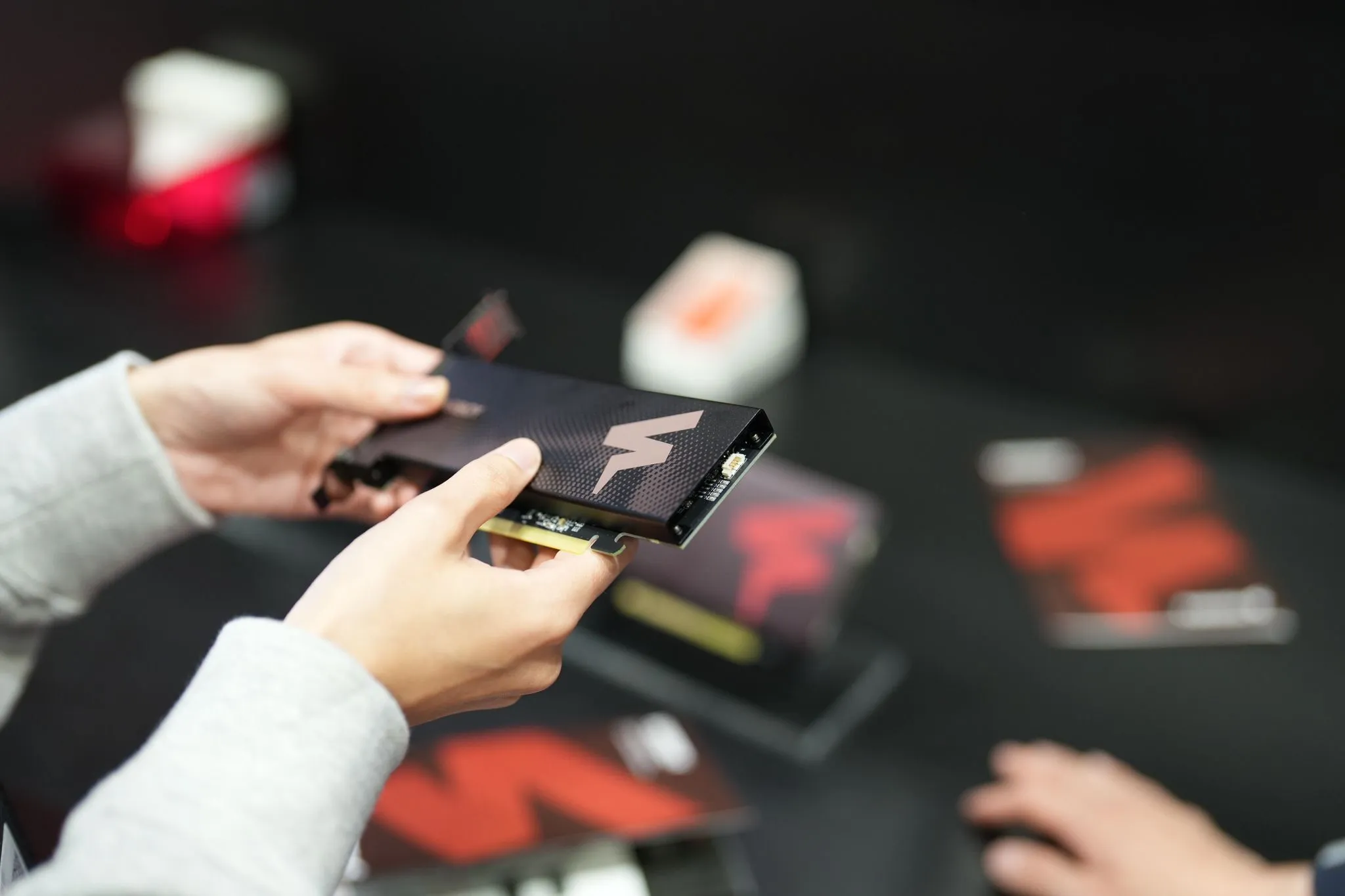 Hands holding a FuriosaAI PCIe accelerator card with a lightning-bolt logo, shown during a hackathon or demo setting, with branded materials and other hardware blurred in the background.