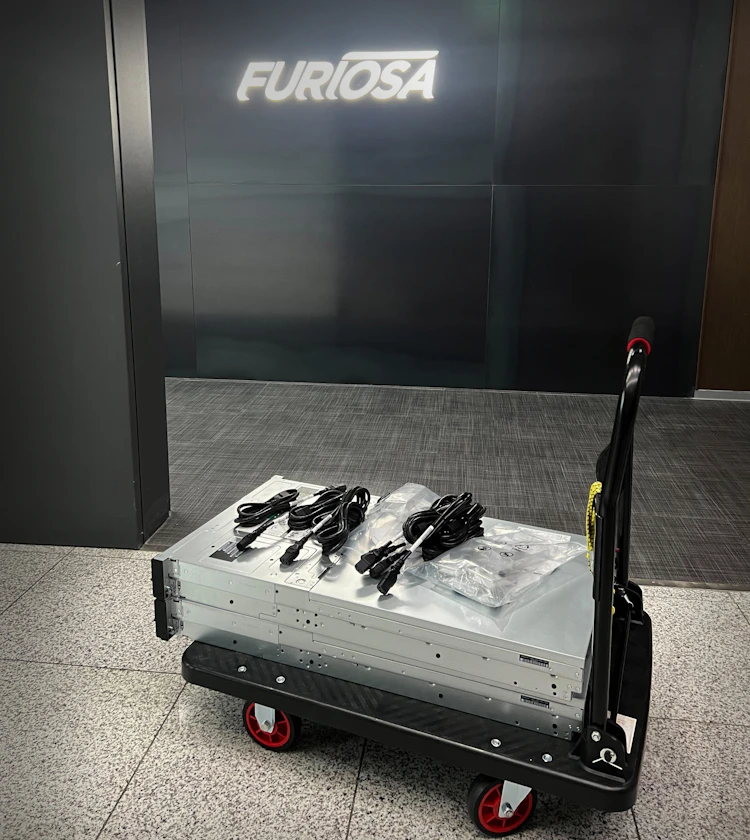 Rack-mounted server placed on a wheeled cart in a hallway with Furiosa branding on the wall behind it, showing power cables and accessories prepared for installation.