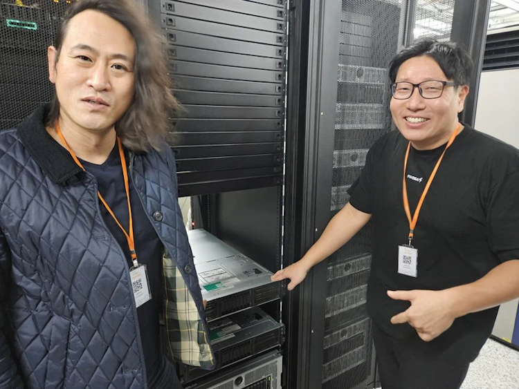 Two people standing beside a server rack in a data center, smiling while presenting rack-mounted servers during a hardware setup or inspection.