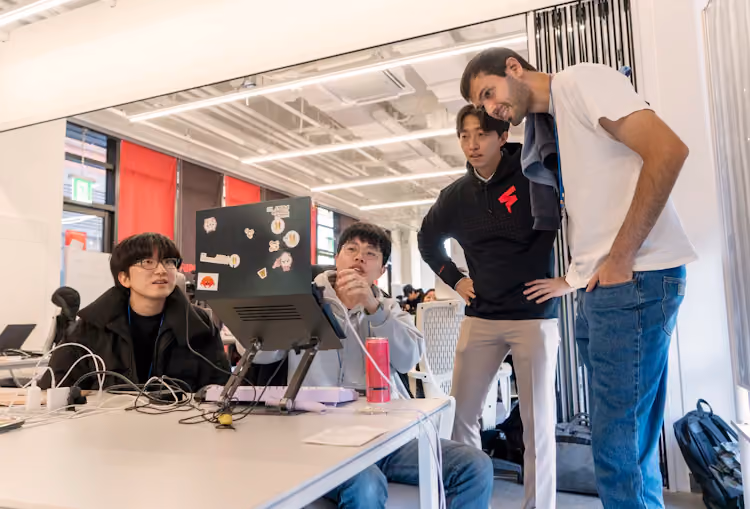 Four participants collaborate at a table during a FuriosaAI hackathon, reviewing code on a laptop with stickers while discussing ideas in a bright, open workspace.