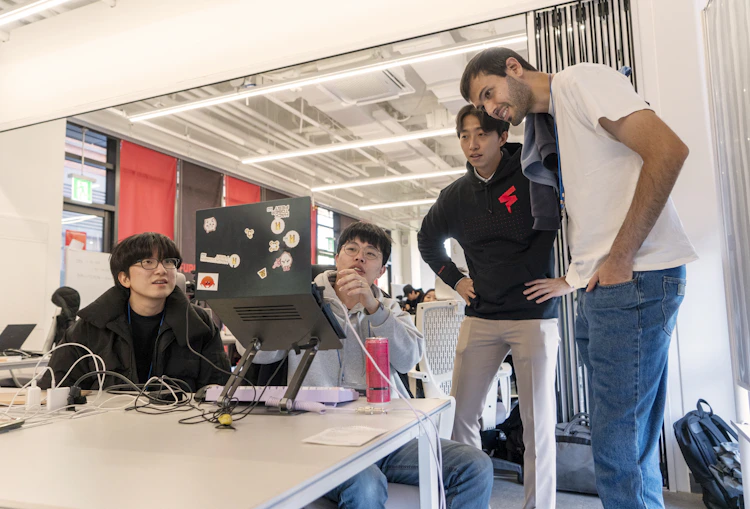 Four participants collaborate at a table during a FuriosaAI hackathon, reviewing code on a laptop with stickers while discussing ideas in a bright, open workspace.