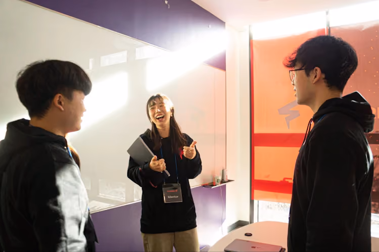 A Furiosa AI hackathon mentor smiles and gestures while talking with two participants in a bright collaboration space, wearing event badges and standing near a whiteboard and laptop.