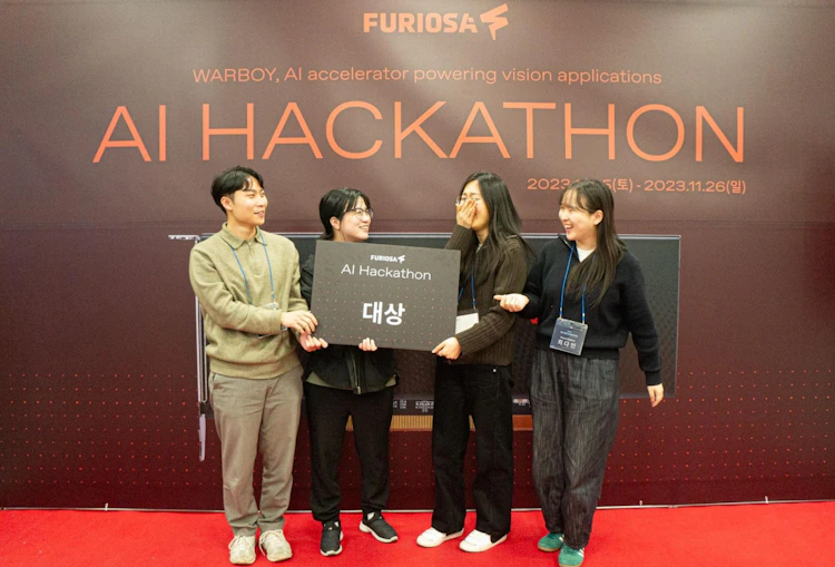 Four participants stand on a stage at the FuriosaAI AI Hackathon, smiling and celebrating while holding a large award sign, with a branded backdrop reading “AI Hackathon” and event dates behind them.