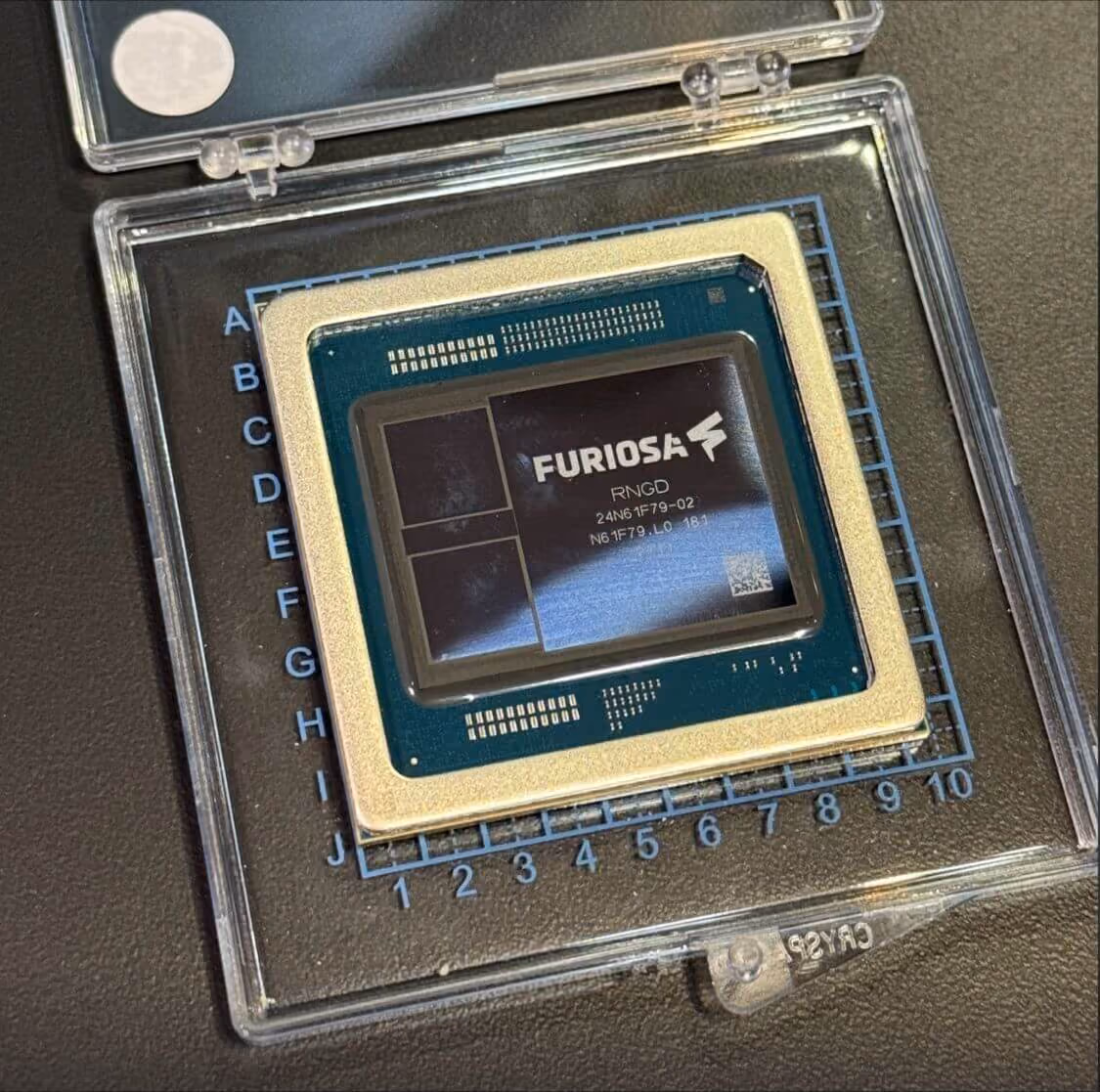 Furiosa RNGD AI processor chip displayed in a transparent protective case, showing the labeled semiconductor package and exposed die resting on a measurement grid.