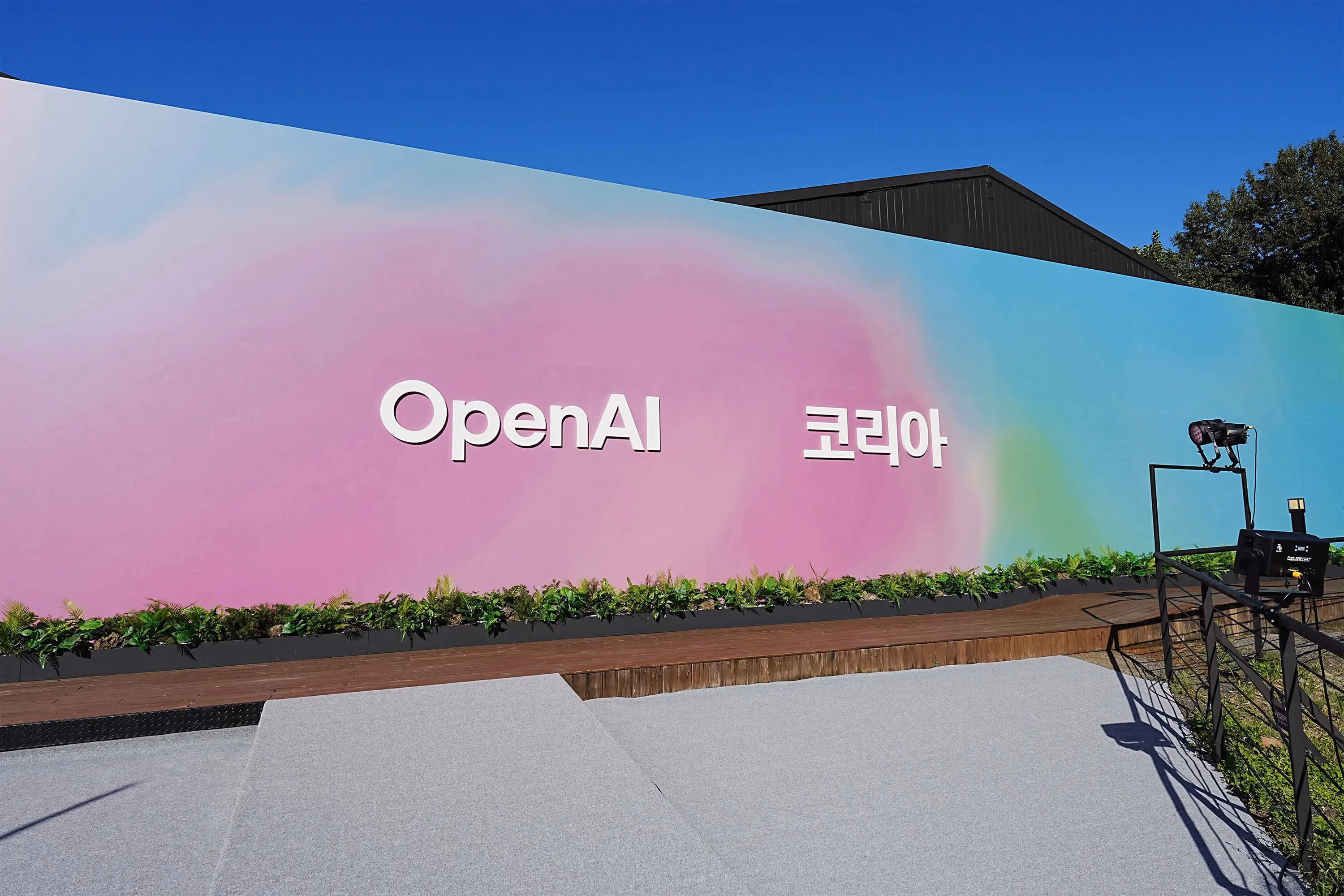 Large outdoor event backdrop with a pastel gradient wall displaying the text “OpenAI Korea,” set against a clear blue sky with a small stage and lighting equipment in front.