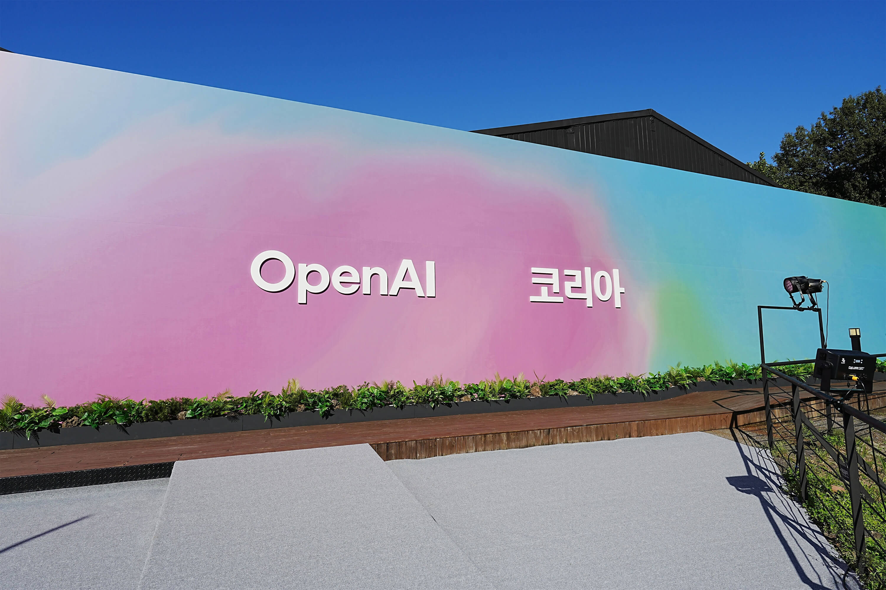 Large outdoor event backdrop with a pastel gradient wall displaying the text “OpenAI Korea,” set against a clear blue sky with a small stage and lighting equipment in front.