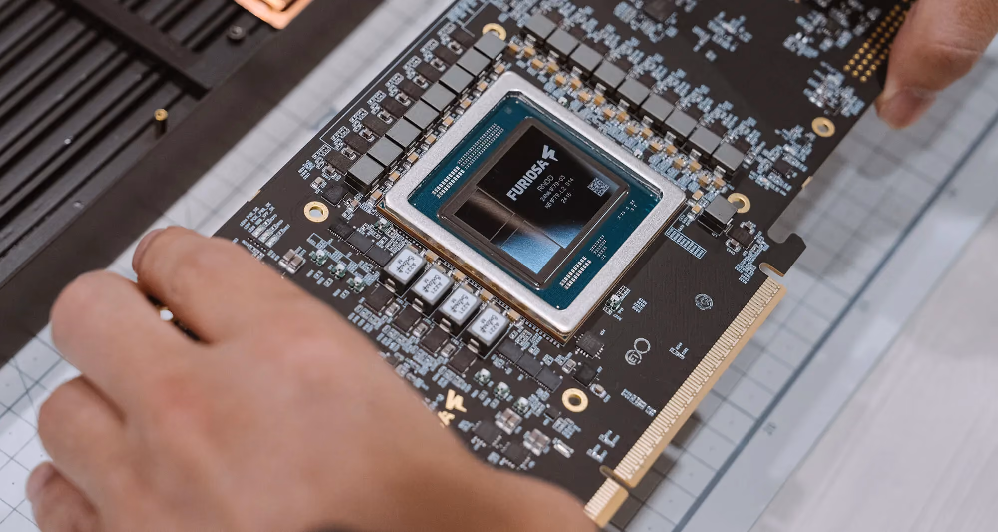 Close-up of a Furiosa RNGD AI accelerator card on a workbench, showing the exposed processor die, surrounding power components, and PCIe edge connector while being handled during inspection or assembly.