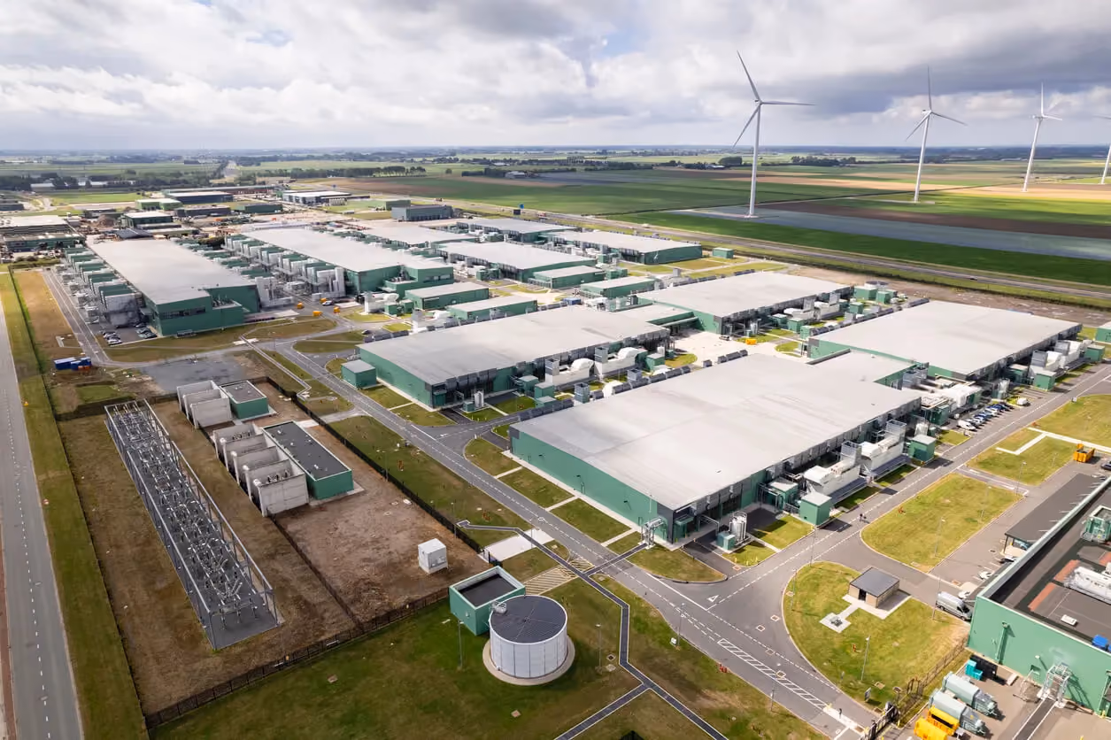 Alt text: Aerial view of a large data center campus with multiple low-profile buildings, surrounded by roads, green fields, and wind turbines, illustrating modern data infrastructure integrated with renewable energy sources.