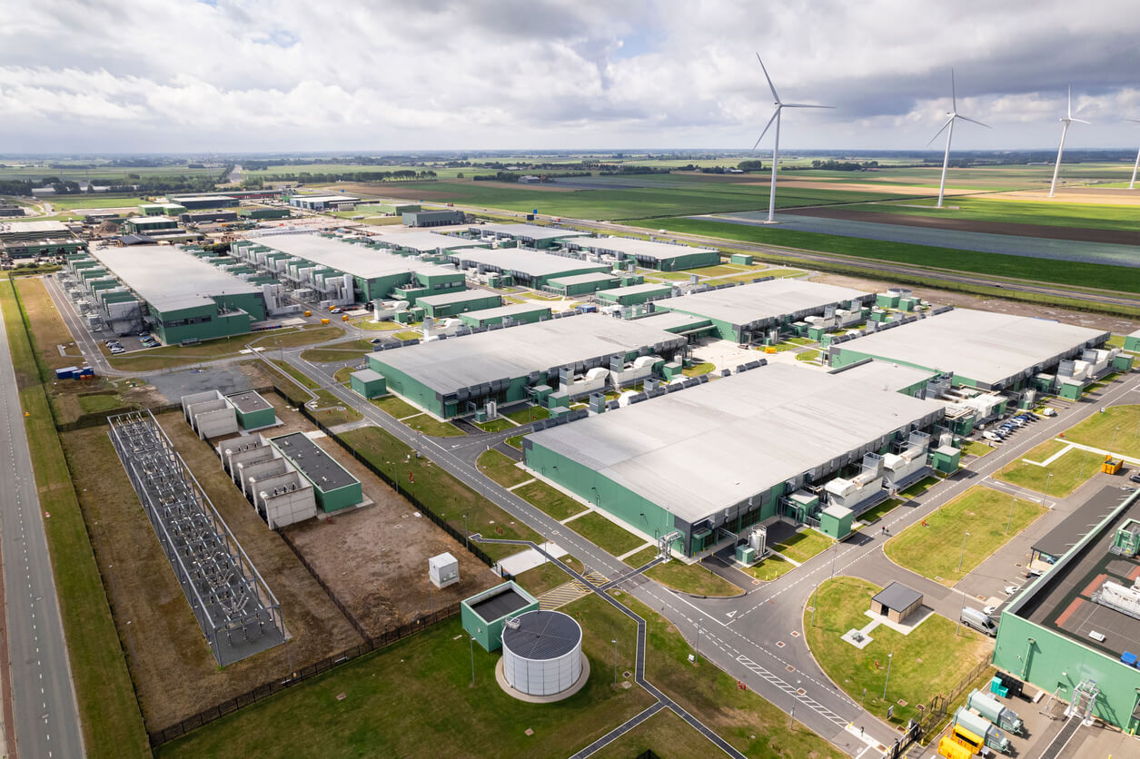 Alt text: Aerial view of a large data center campus with multiple low-profile buildings, surrounded by roads, green fields, and wind turbines, illustrating modern data infrastructure integrated with renewable energy sources.