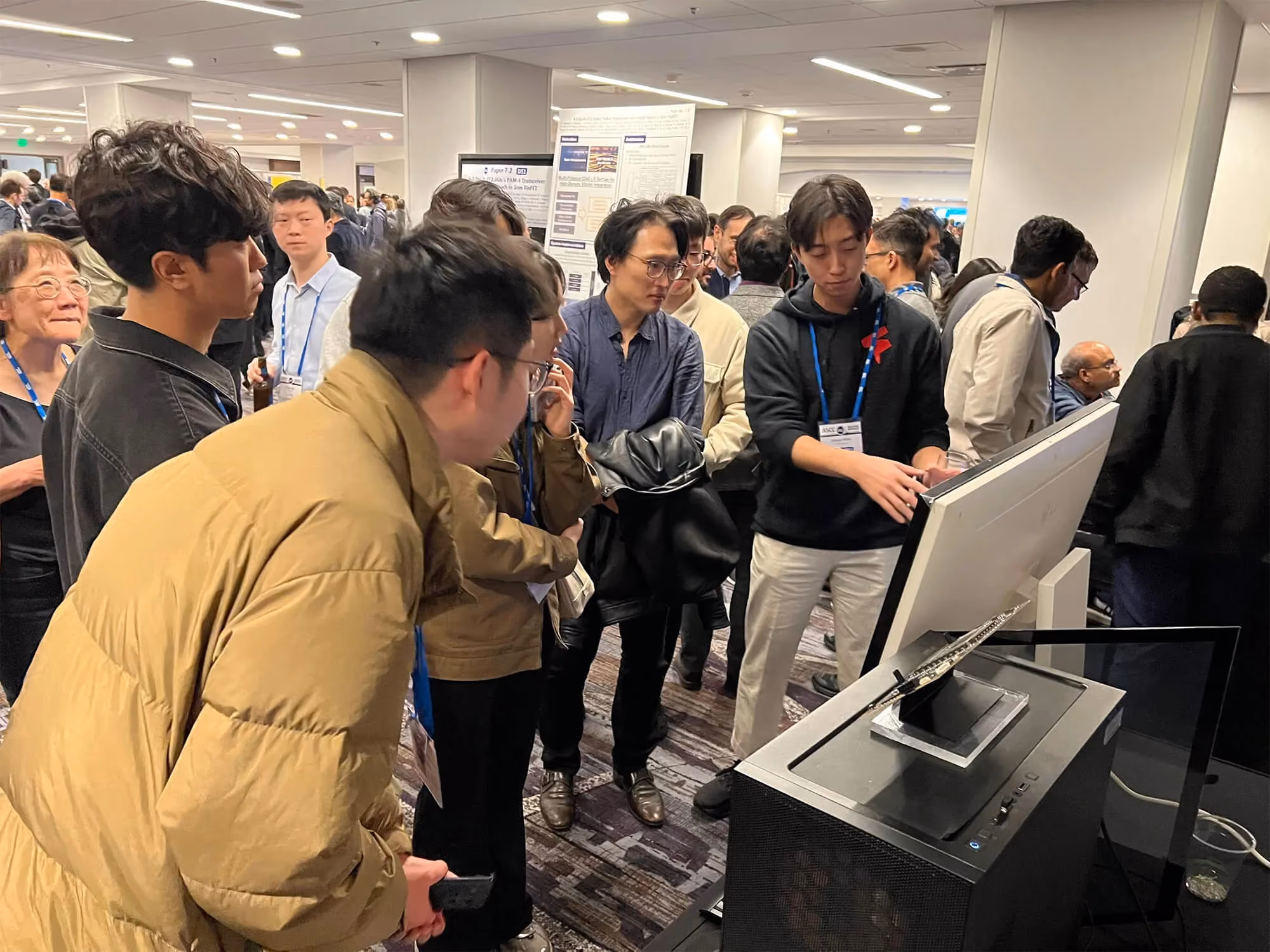 Conference attendees gathered around a live Furiosa hardware and AI inference demo, watching a presenter explain the system at an industry event floor with posters and booths in the background.