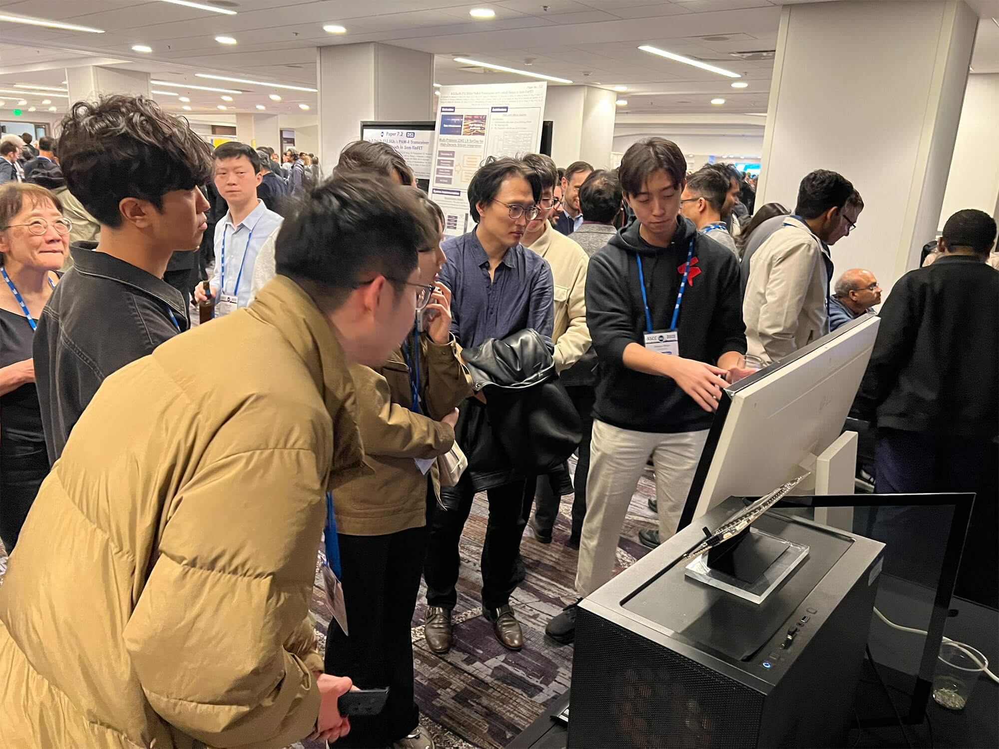 Conference attendees gathered around a live Furiosa hardware and AI inference demo, watching a presenter explain the system at an industry event floor with posters and booths in the background.