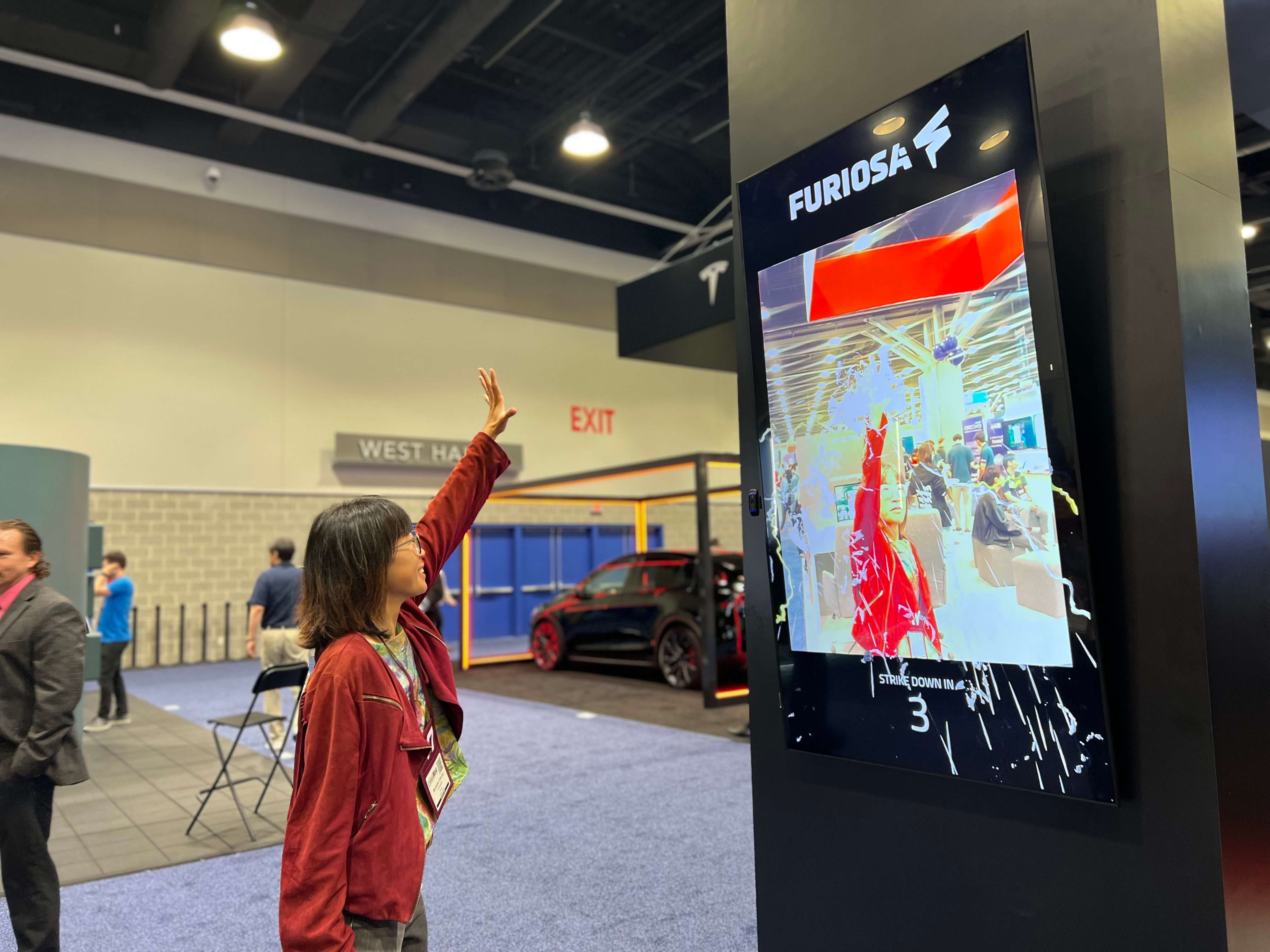 Person interacting with a FuriosaAI demo at a tech conference booth, raising their hand toward a tall digital display labeled “FURIOSA.” The screen shows a real-time computer vision application tracking the raised arm with visual effects, while other attendees and exhibition booths are visible in the background.