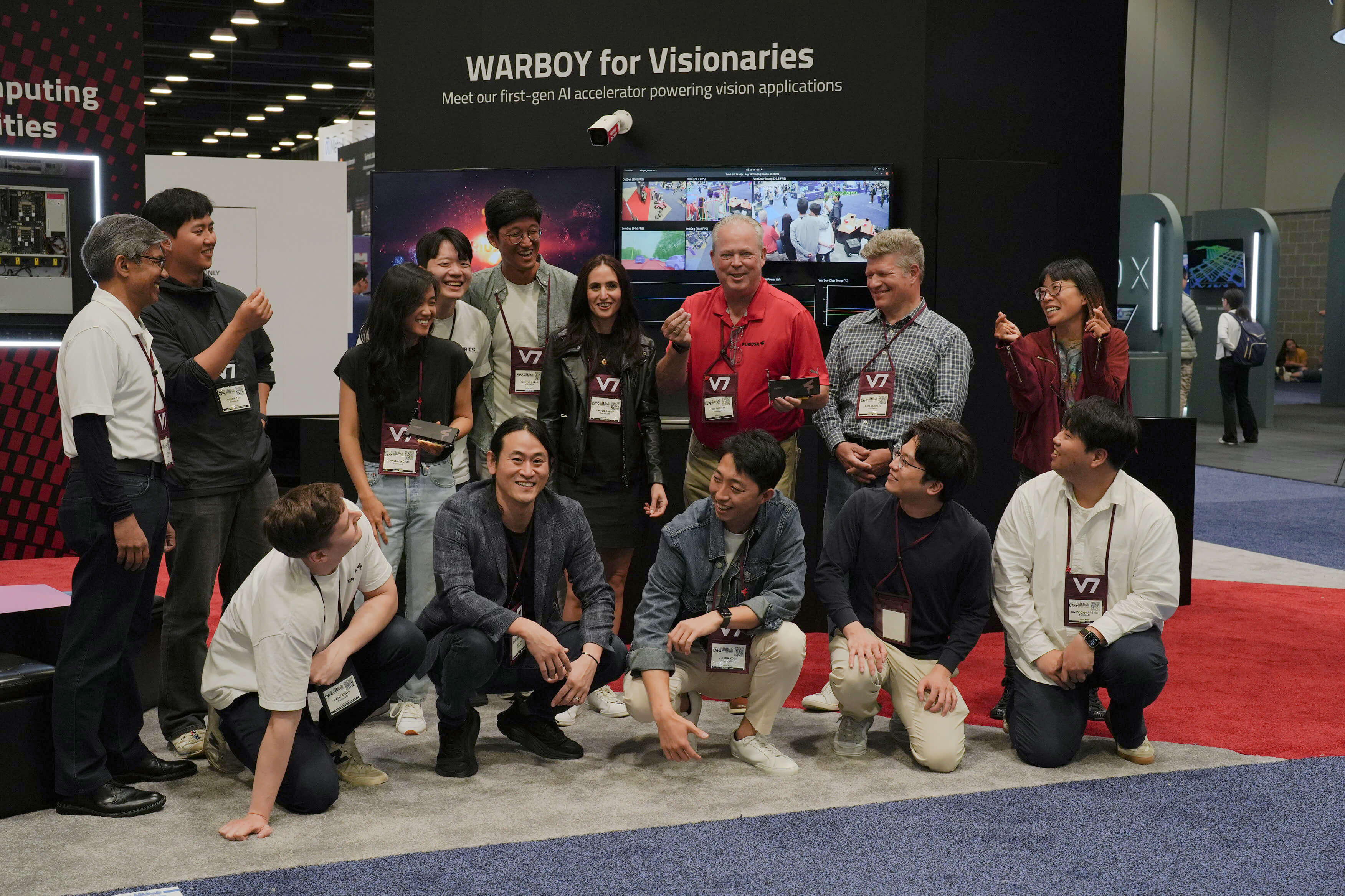 Group photo at a technology conference booth labeled “WARBOY for Visionaries,” showing a diverse team of attendees and staff smiling and posing together in front of large screens demonstrating computer vision applications. Several people wear conference badges and hold small hardware cards, capturing a celebratory moment around the AI accelerator showcase.