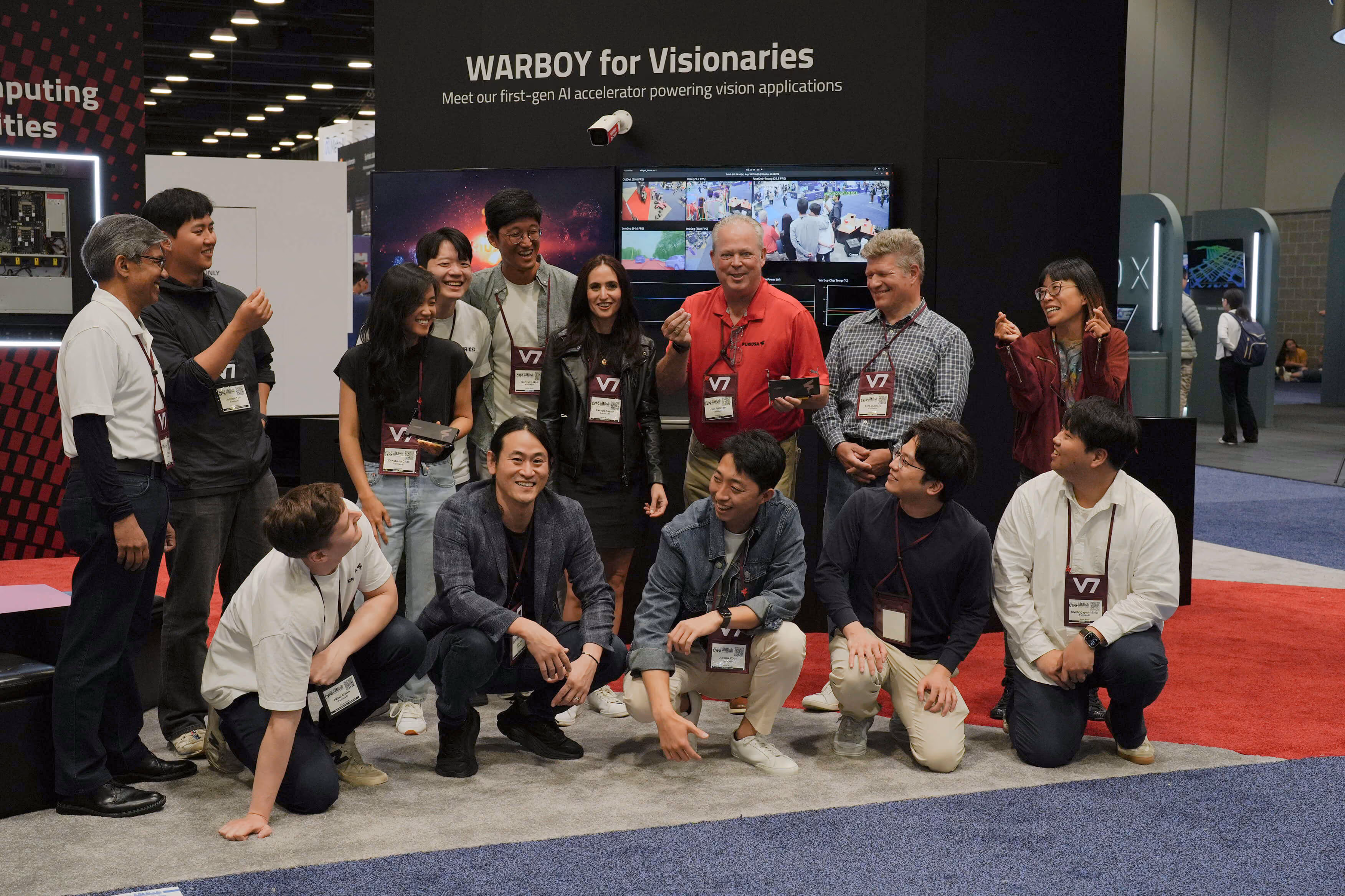 Group photo at a technology conference booth labeled “WARBOY for Visionaries,” showing a diverse team of attendees and staff smiling and posing together in front of large screens demonstrating computer vision applications. Several people wear conference badges and hold small hardware cards, capturing a celebratory moment around the AI accelerator showcase.