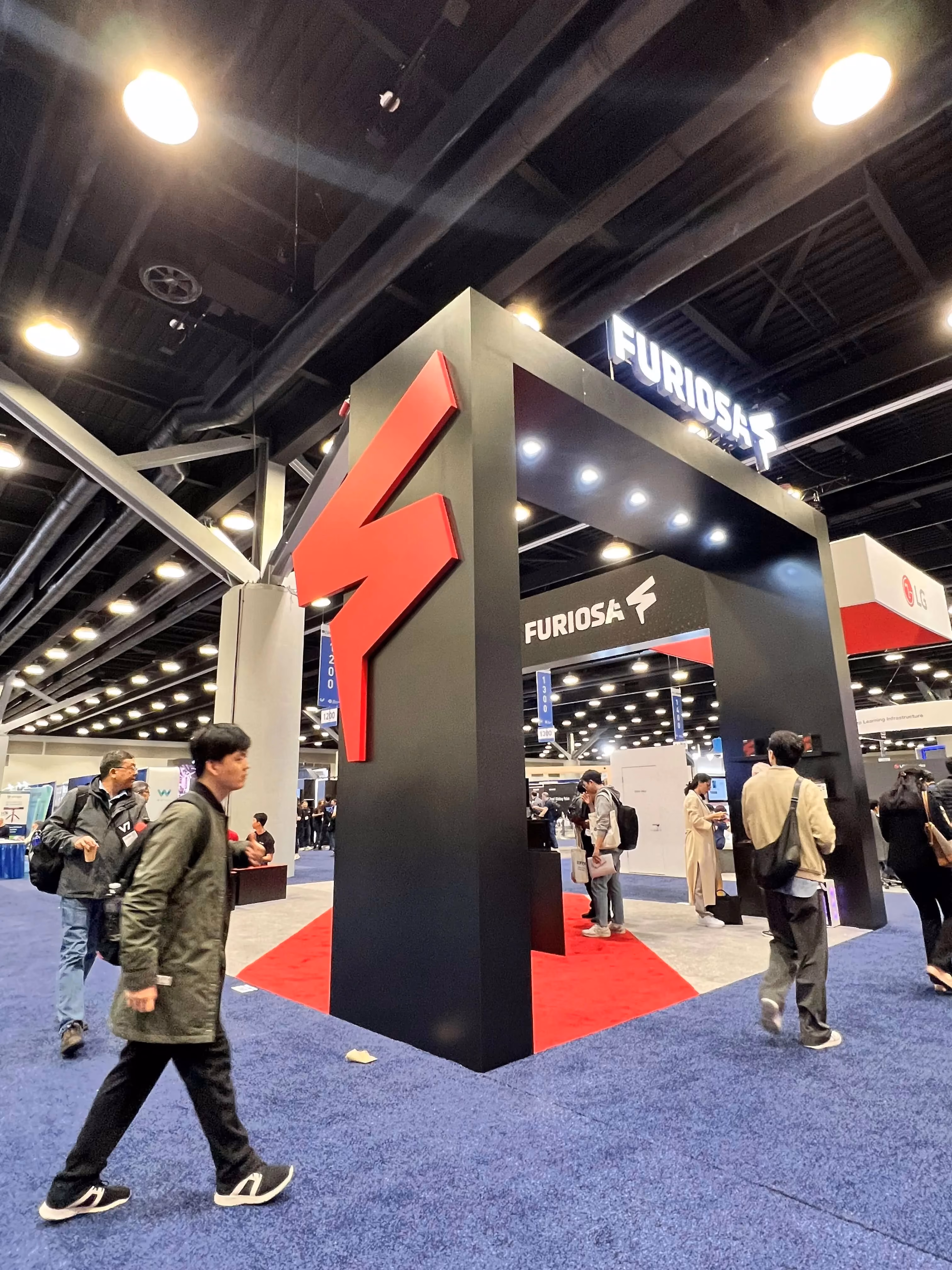 Wide-angle view of the FuriosaAI exhibition booth at a large technology conference, featuring a tall black structure with a bold red lightning-bolt logo and illuminated “FURIOSA” signage overhead. Attendees walk past and gather around the booth on a red-and-blue carpeted floor, highlighting the company’s prominent presence on the show floor.