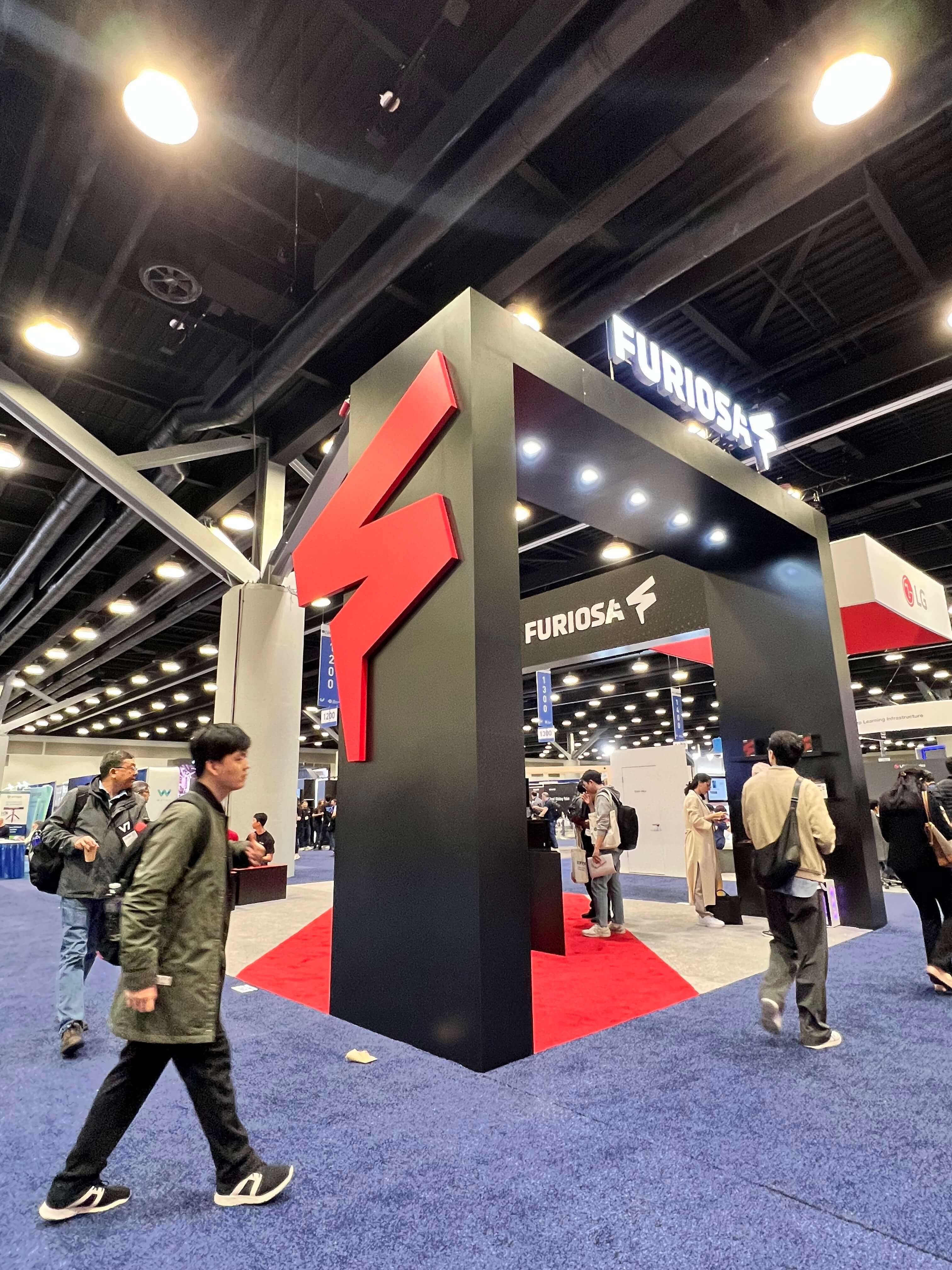 Wide-angle view of the FuriosaAI exhibition booth at a large technology conference, featuring a tall black structure with a bold red lightning-bolt logo and illuminated “FURIOSA” signage overhead. Attendees walk past and gather around the booth on a red-and-blue carpeted floor, highlighting the company’s prominent presence on the show floor.