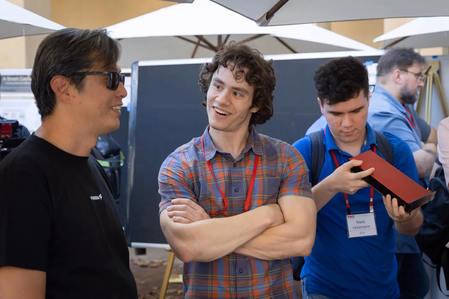 Three attendees at a tech conference booth chat and examine hardware, with one person smiling and crossing their arms, another wearing sunglasses, and a third inspecting a red device, all wearing event badges in an outdoor expo setting.