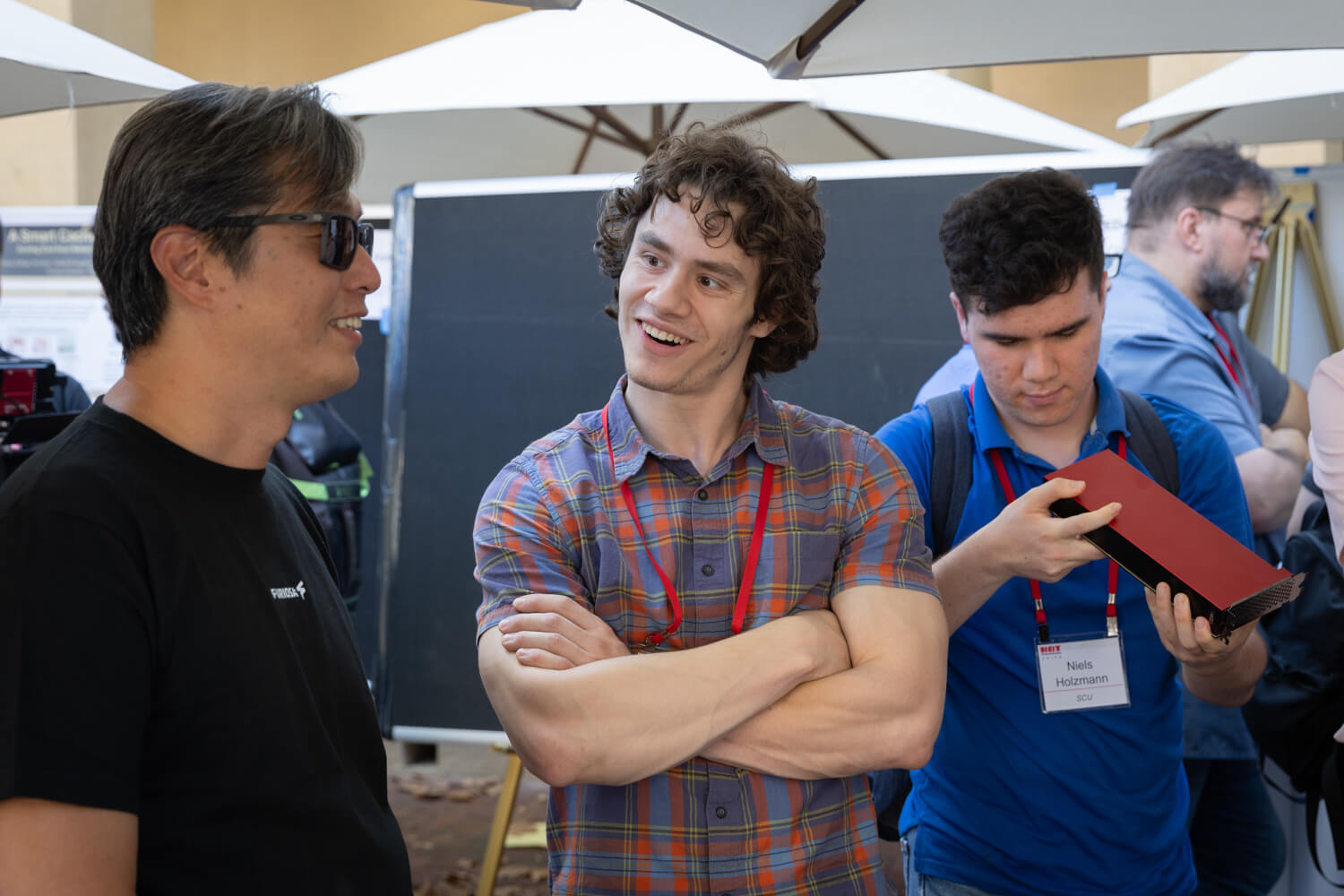 Three attendees at a tech conference booth chat and examine hardware, with one person smiling and crossing their arms, another wearing sunglasses, and a third inspecting a red device, all wearing event badges in an outdoor expo setting.