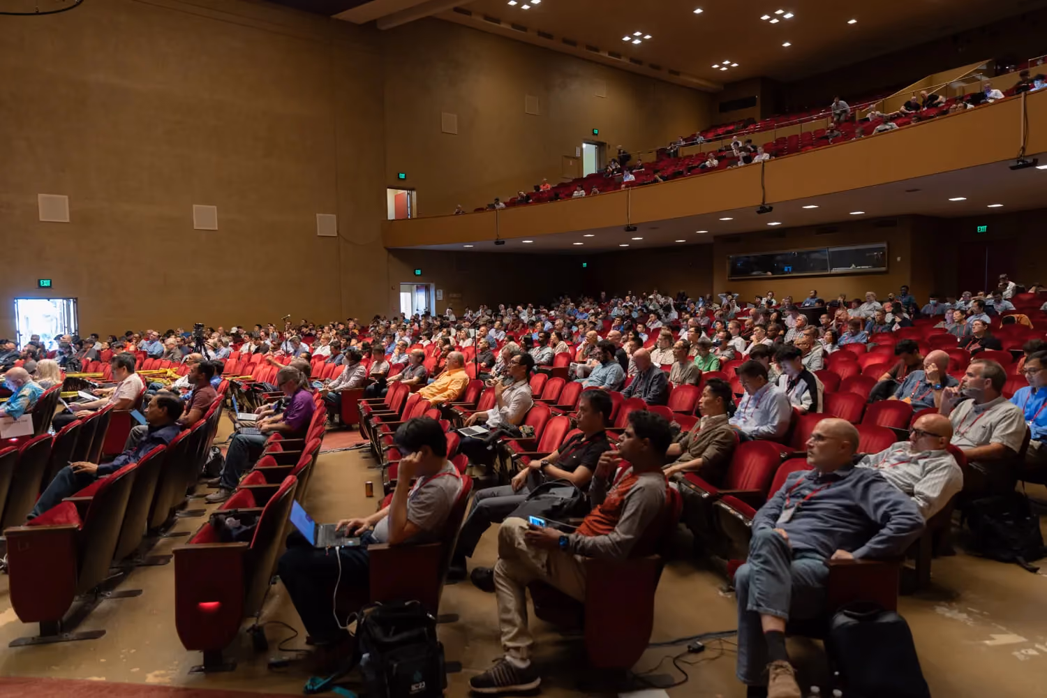 Wide view of a large auditorium filled with attendees seated in red chairs, watching a technical conference presentation, with audience members using laptops and devices during a session at an industry event such as Hot Chips.