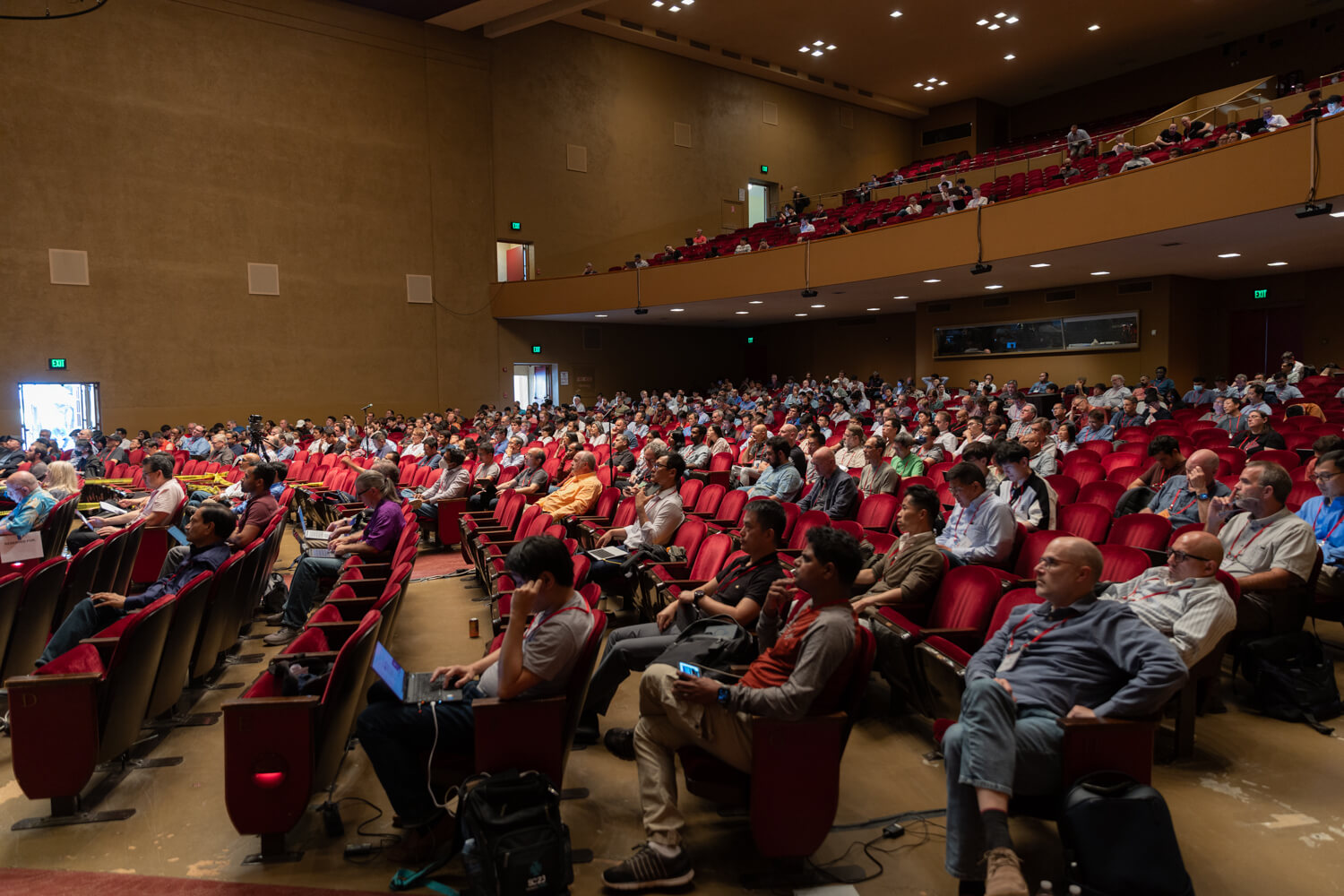 Wide view of a large auditorium filled with attendees seated in red chairs, watching a technical conference presentation, with audience members using laptops and devices during a session at an industry event such as Hot Chips.