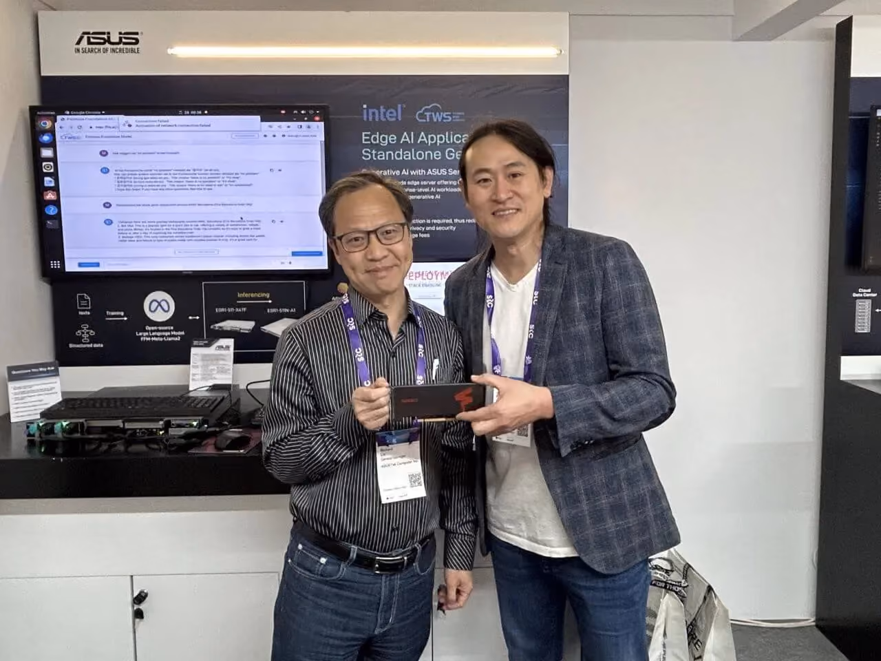 Two men standing at an ASUS exhibition booth, smiling while holding a FuriosaAI accelerator card. Behind them, a display screen shows an edge AI application demo with Intel and ASUS branding, indicating a collaborative technology showcase.