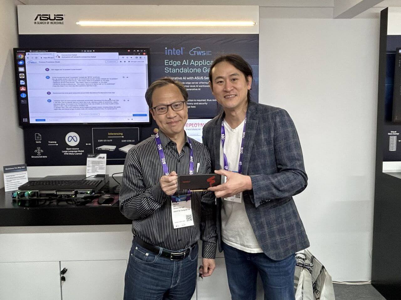 Two men standing at an ASUS exhibition booth, smiling while holding a FuriosaAI accelerator card. Behind them, a display screen shows an edge AI application demo with Intel and ASUS branding, indicating a collaborative technology showcase.