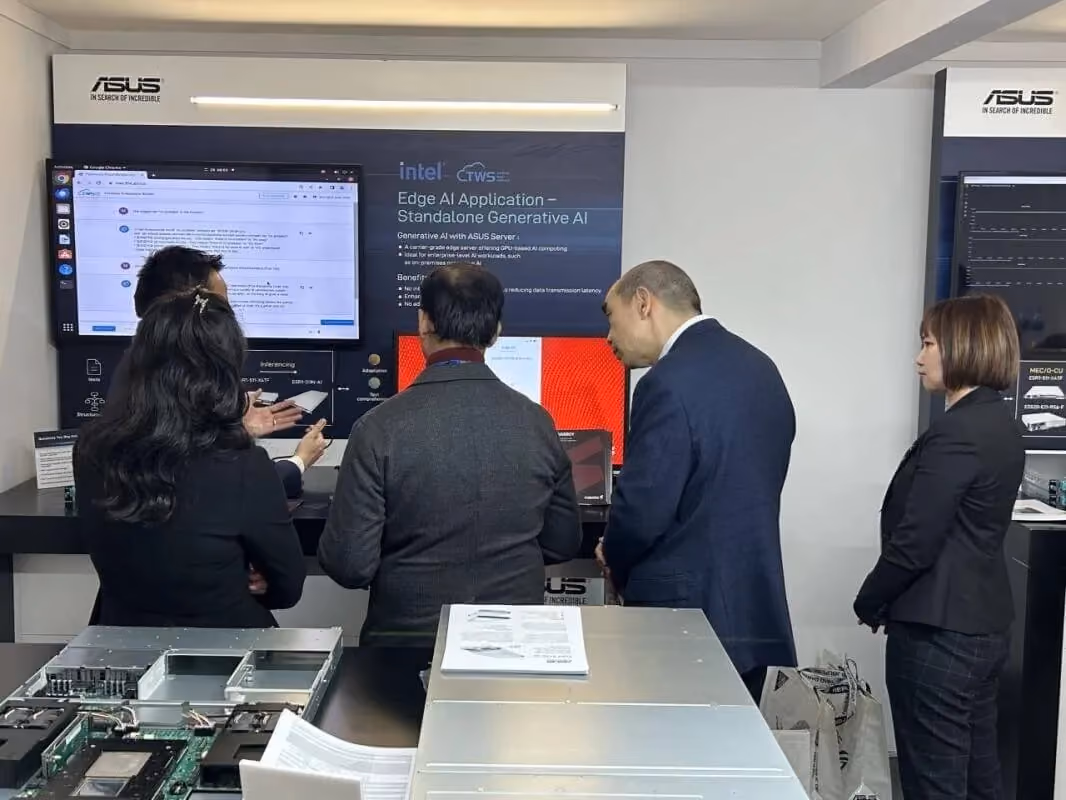 A small group of professionals gathered at an ASUS demo booth, viewed from behind, watching a live demonstration of an edge AI and standalone generative AI application. Large display screens show technical interfaces and Intel–ASUS branding, while server hardware and documentation are visible on the counter in the foreground.