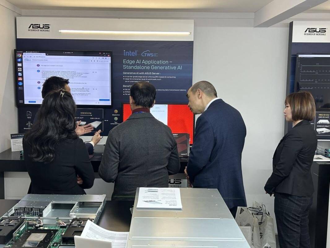 A small group of professionals gathered at an ASUS demo booth, viewed from behind, watching a live demonstration of an edge AI and standalone generative AI application. Large display screens show technical interfaces and Intel–ASUS branding, while server hardware and documentation are visible on the counter in the foreground.