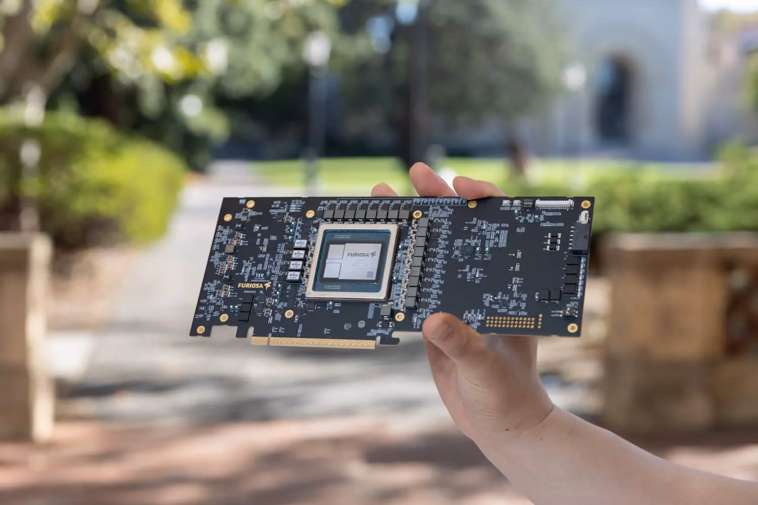 Hand holding a Furiosa RNGD NPU accelerator card outdoors, showing the processor chip and surrounding circuitry.