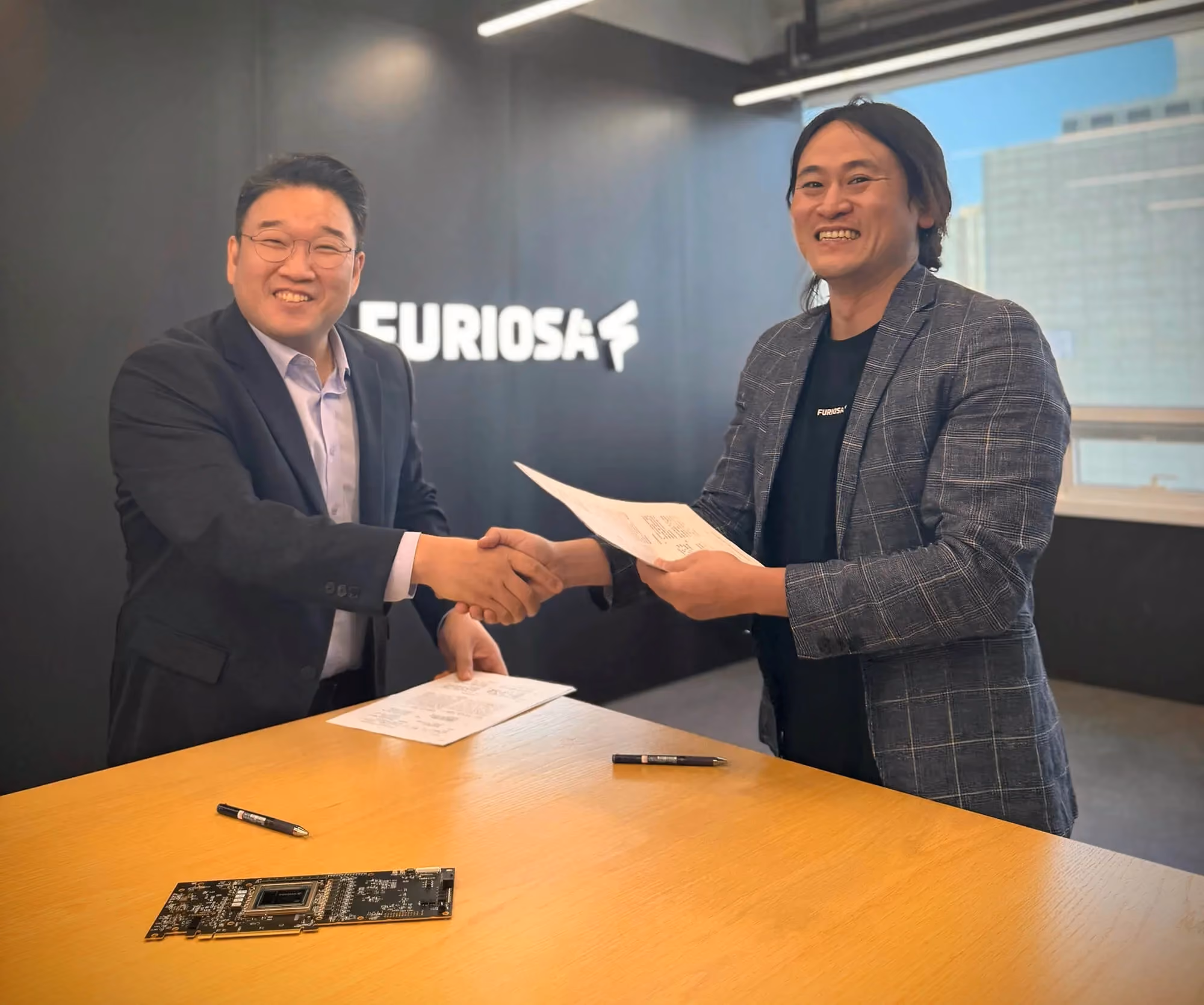 Two business leaders shaking hands while exchanging signed documents at a table inside the FuriosaAI office.