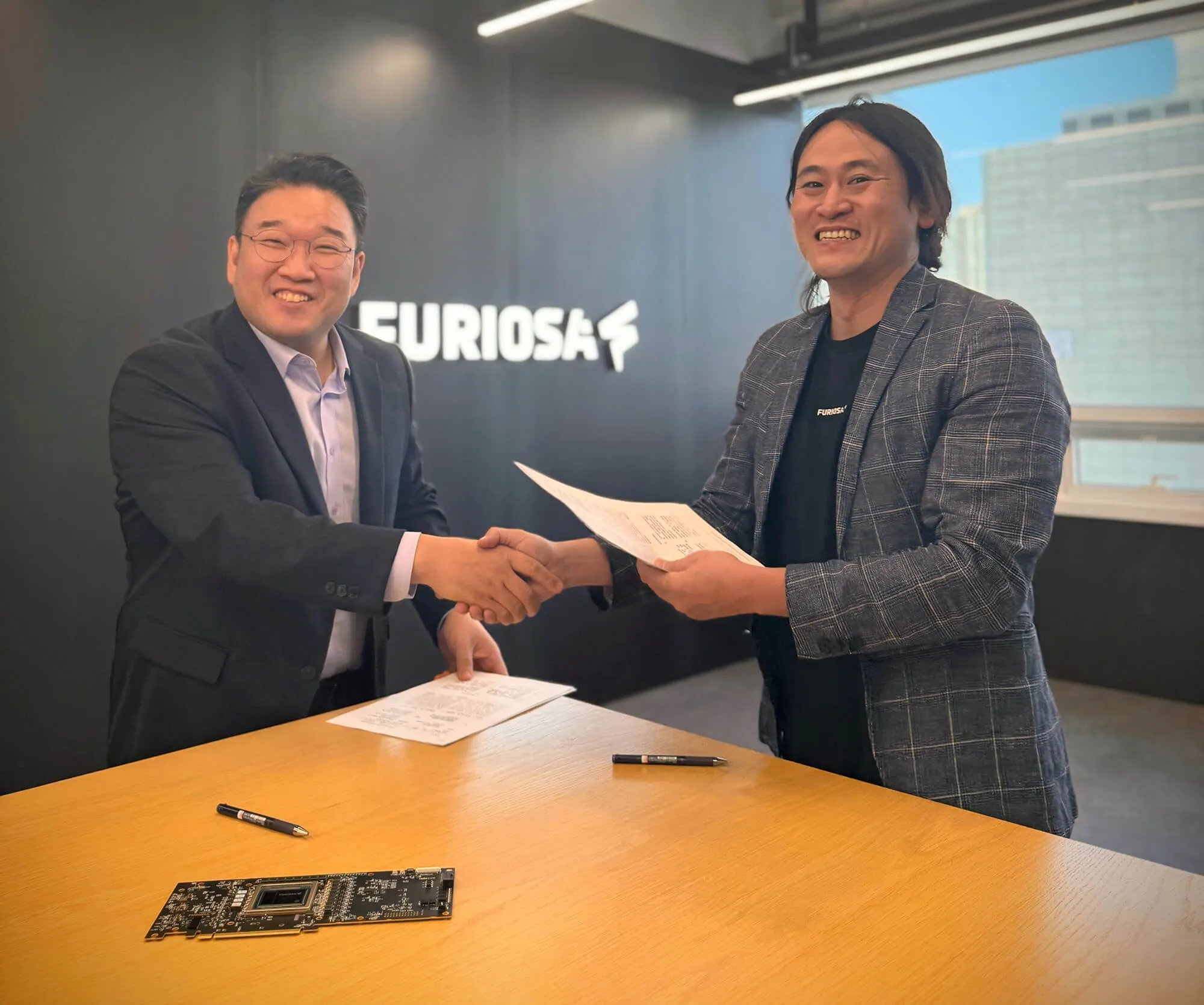 Two business leaders shaking hands while exchanging signed documents at a table inside the FuriosaAI office.