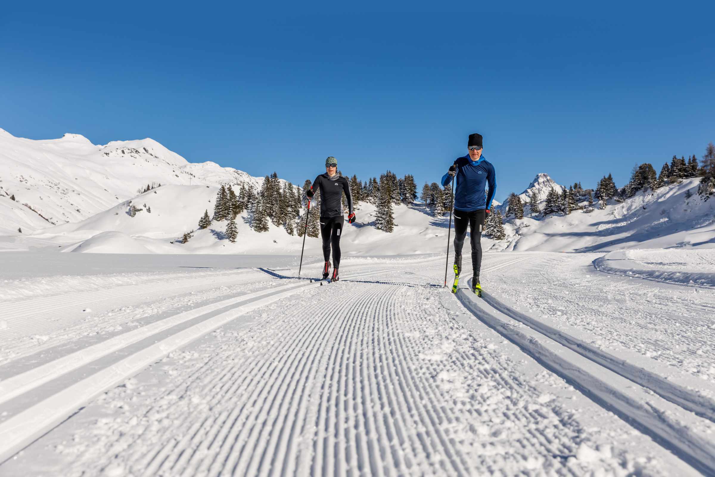 Zwei Langläufer in schwarzer und blauer Kleidung skaten auf gespurter Loipe in schneebedeckter Berglandschaft bei klarem blauem Himmel.