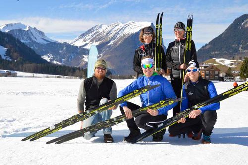 Fünf Skifahrer mit Langlaufski posieren lachend auf schneebedecktem Feld vor schneebedeckten Bergen bei klarem Himmel.