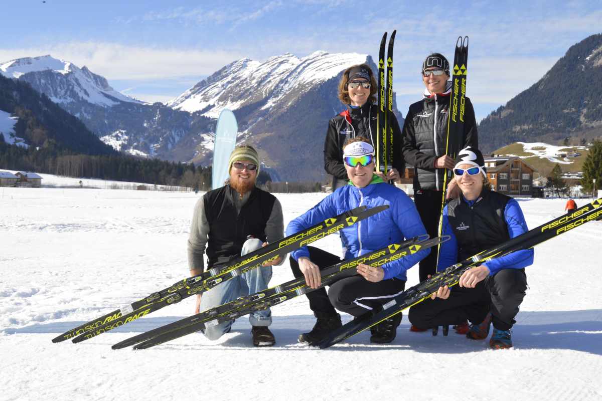 Fünf Skifahrer mit Langlaufski posieren lachend auf schneebedecktem Feld vor schneebedeckten Bergen bei klarem Himmel.