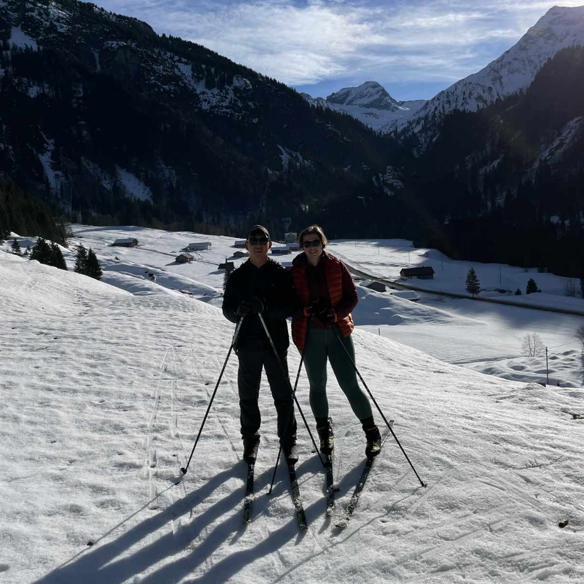 Zwei Menschen beim Skifahren auf einer schneebedeckten Landschaft mit Bergen und Hütten im Hintergrund.