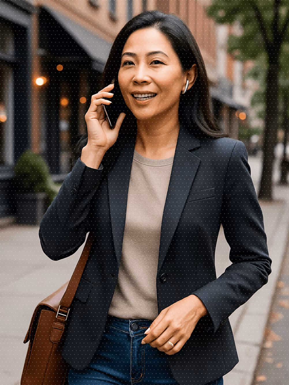 Smiling woman in black blazer and beige top talking on a smartphone while walking outdoors with a brown shoulder bag.