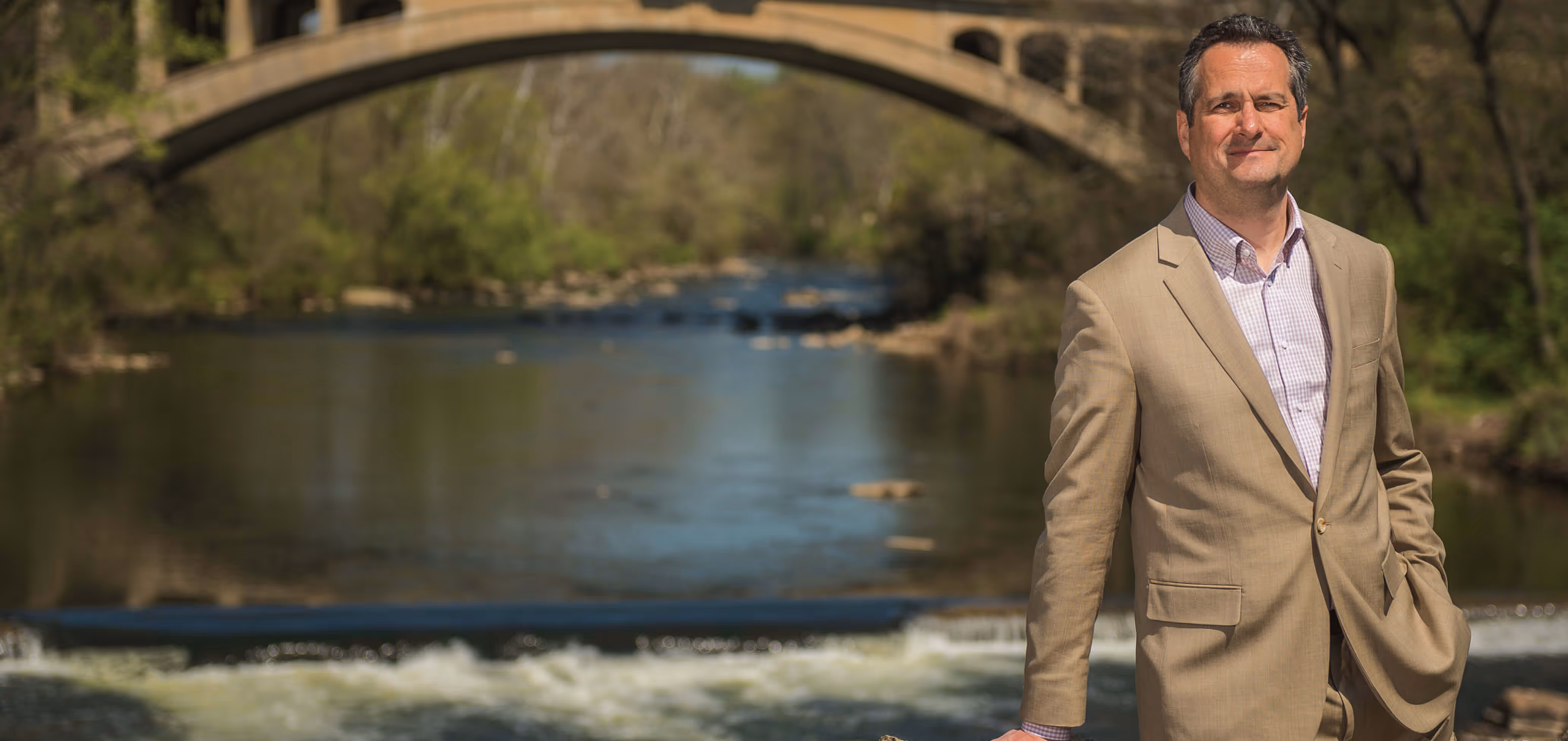 James H. Lee in beige suit standing near a river with a stone arch bridge in the background.