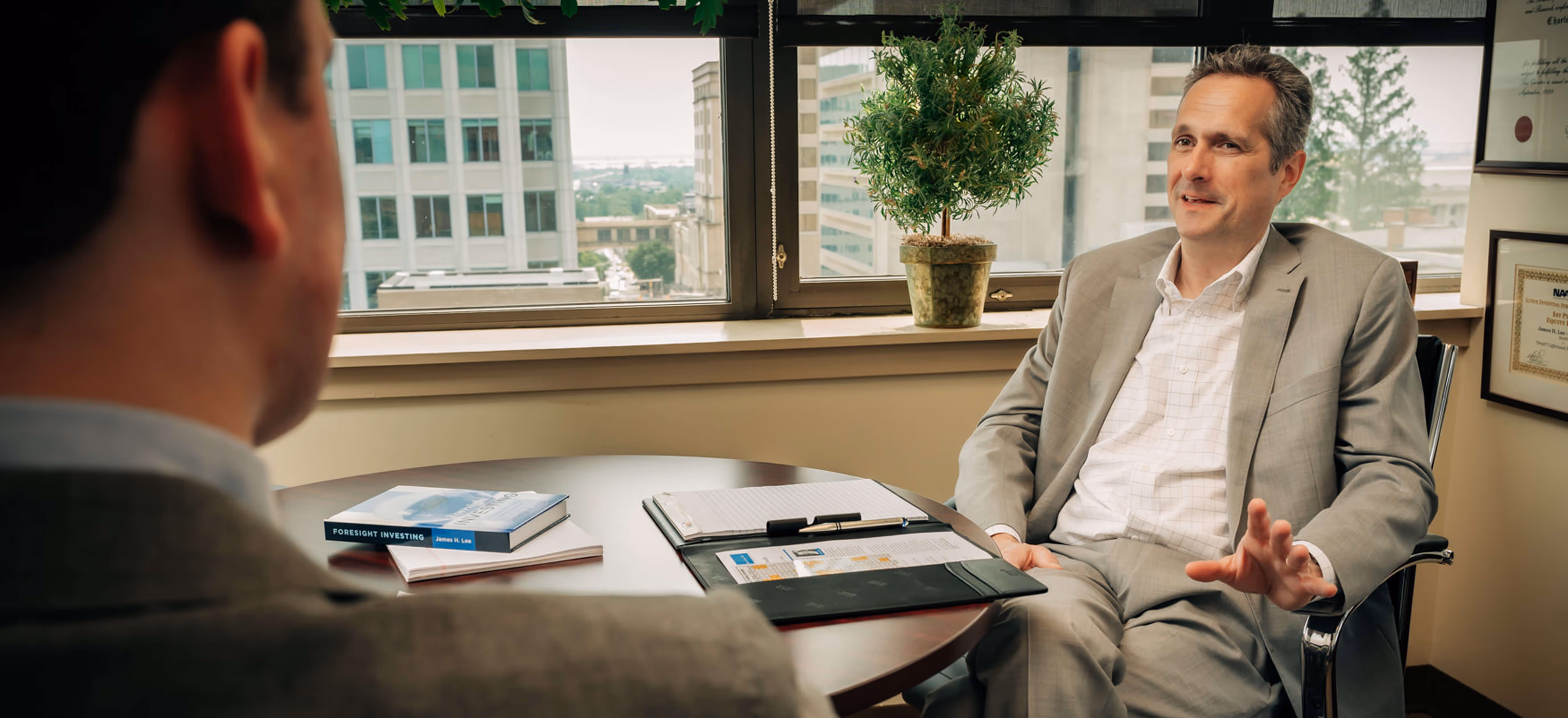 James H. Lee and a man having a discussion in a modern office with a city view through large windows.