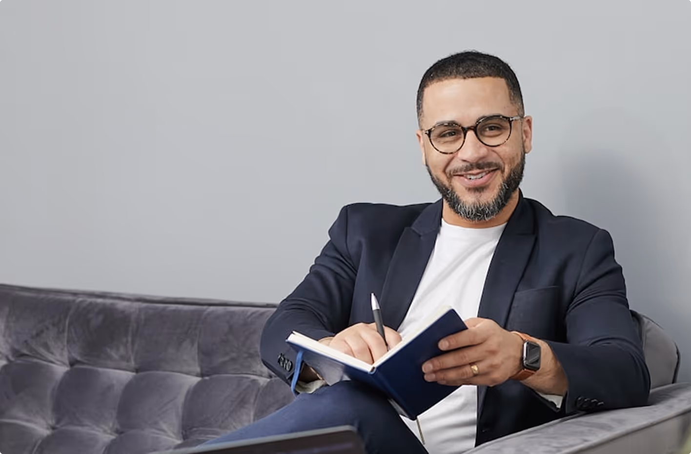 Smiling man with glasses and beard sitting on a gray sofa, writing in a blue notebook.